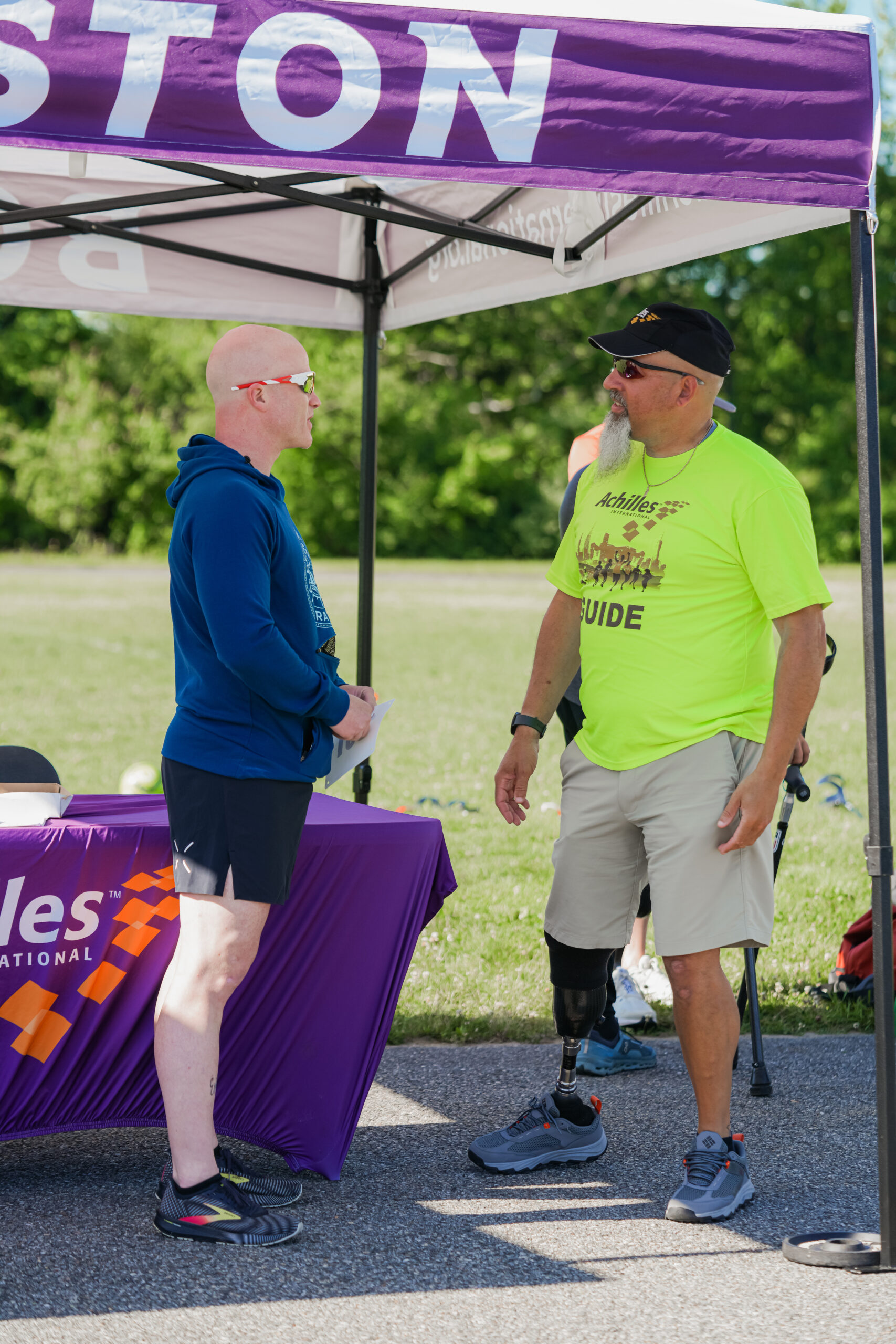 A bald man in a blue hoodie and black shorts talks to a guide wearing a neon yellow 'Achilles Guide' shirt, beige shorts, and a black cap under a tent labeled 'Boston.' The guide has a prosthetic leg and is smiling during their conversation. A purple table with the 'Achilles International' logo is nearby, with other participants visible in the background at the outdoor event.