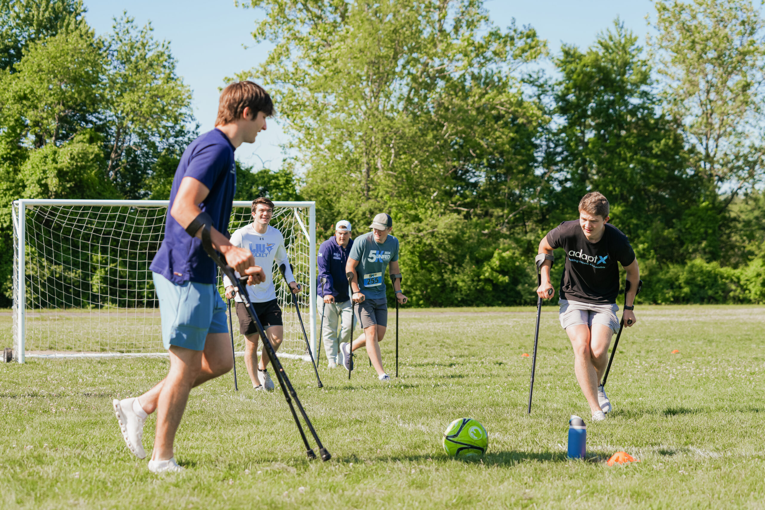 Two athletes using forearm crutches engage in an adaptive soccer game on a grassy field, focusing on a green soccer ball. One wears a navy blue shirt and light blue shorts, while the other wears a black 'adaptX' shirt and gray shorts. Behind them, three other participants with crutches walk toward the game, smiling. A soccer goal and lush green trees are visible in the background.