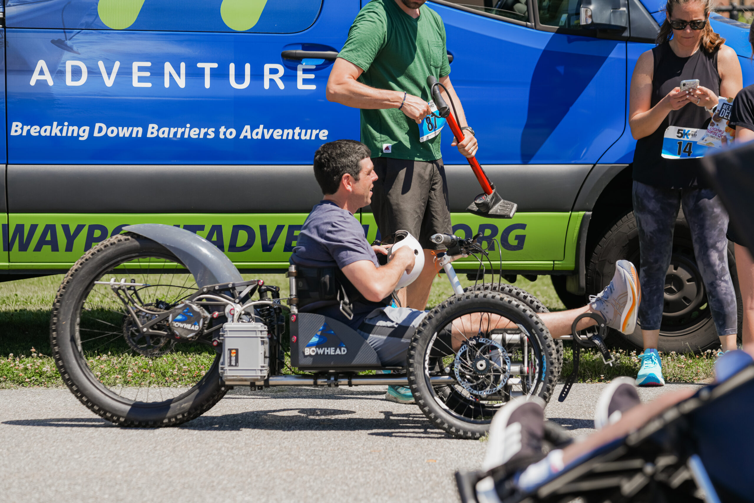 A man with short dark hair and a smile sits in a Bowhead adaptive off-road handcycle, gripping the handlebars. He is wearing a gray T-shirt, shorts, and white sneakers with orange accents. The handcycle has rugged tires and an electric assist system. Behind him, a blue and green van with the words "Breaking Down Barriers to Adventure" is visible. A person in a green shirt holds a red mobility cane, while another participant in athletic gear checks their phone. The setting is an outdoor event focused on adaptive sports and inclusivity.