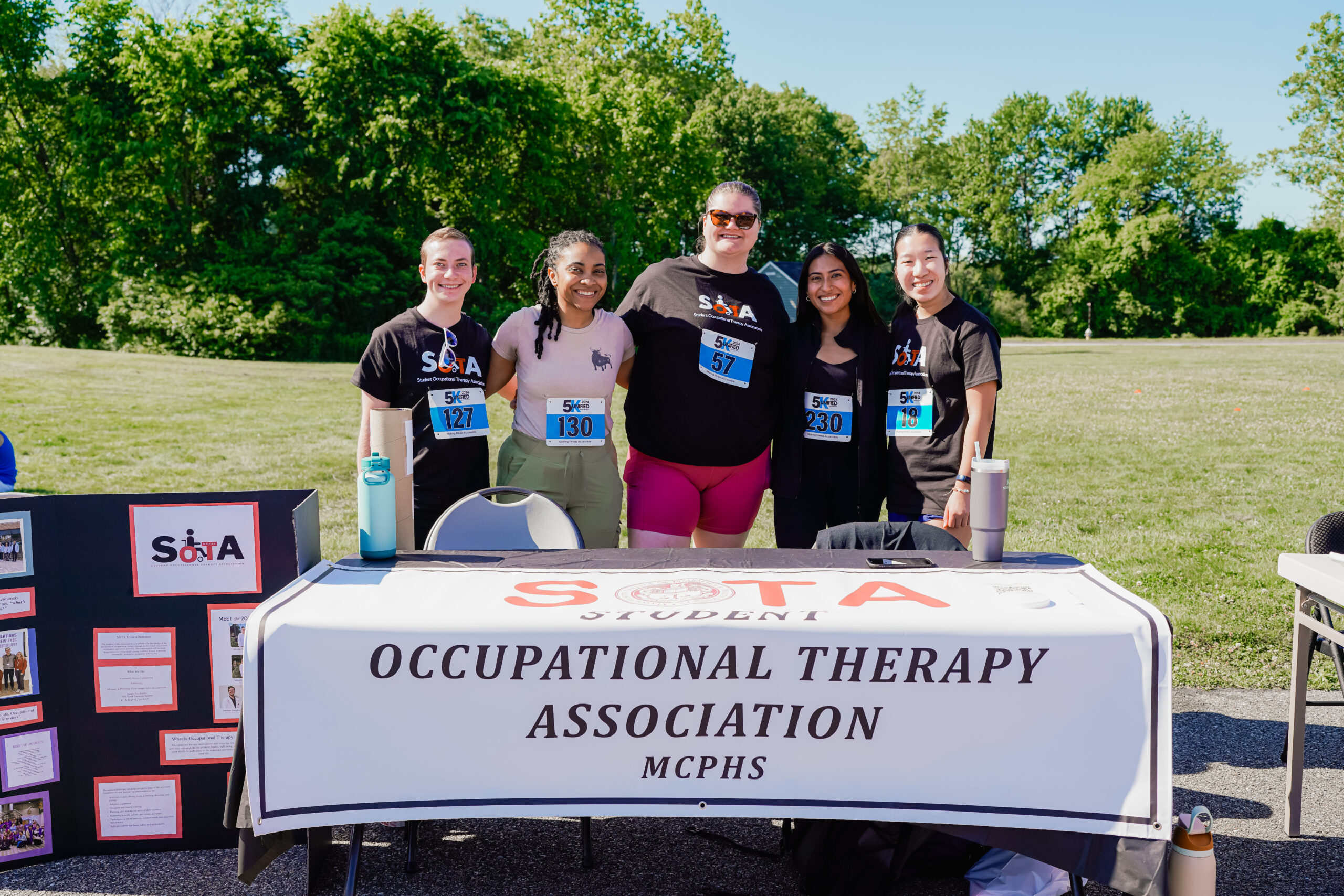 Five individuals stand together, smiling behind a booth for the Student Occupational Therapy Association (SOTA) from MCPHS. Three of them wear black SOTA shirts, while two wear casual athletic attire, all with race bibs. The table is covered with a white banner displaying the organization's name, and to the left, an informational board with photos and text about the group is set up. Water bottles and other small items are arranged on the table. The background features a grassy field with trees and a clear blue sky.