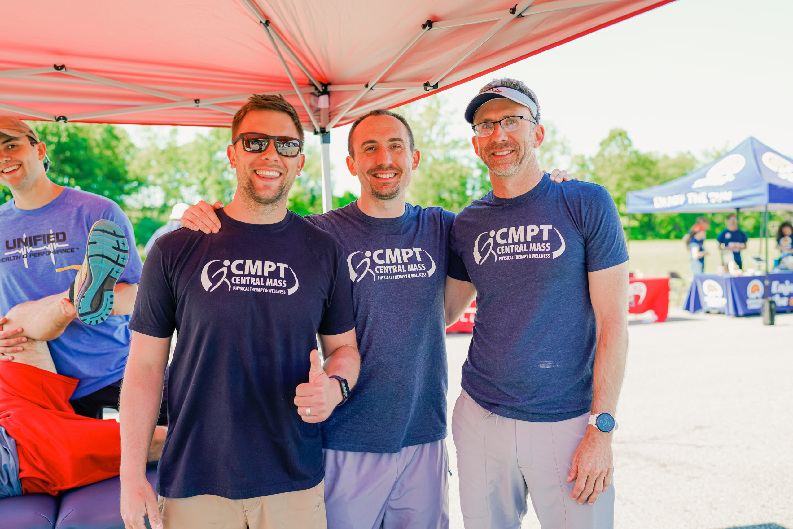 Three men wearing matching navy blue "CMPT Central Mass Physical Therapy & Wellness" t-shirts stand together under a red canopy tent, smiling at the camera. The man on the left is wearing sunglasses and giving a thumbs-up, while the other two have their arms around each other. In the background, a person in a blue "Unified Health & Performance" shirt is lying on a table with their leg stretched, and event tents with banners and people are visible. The setting is outdoors on a sunny day.