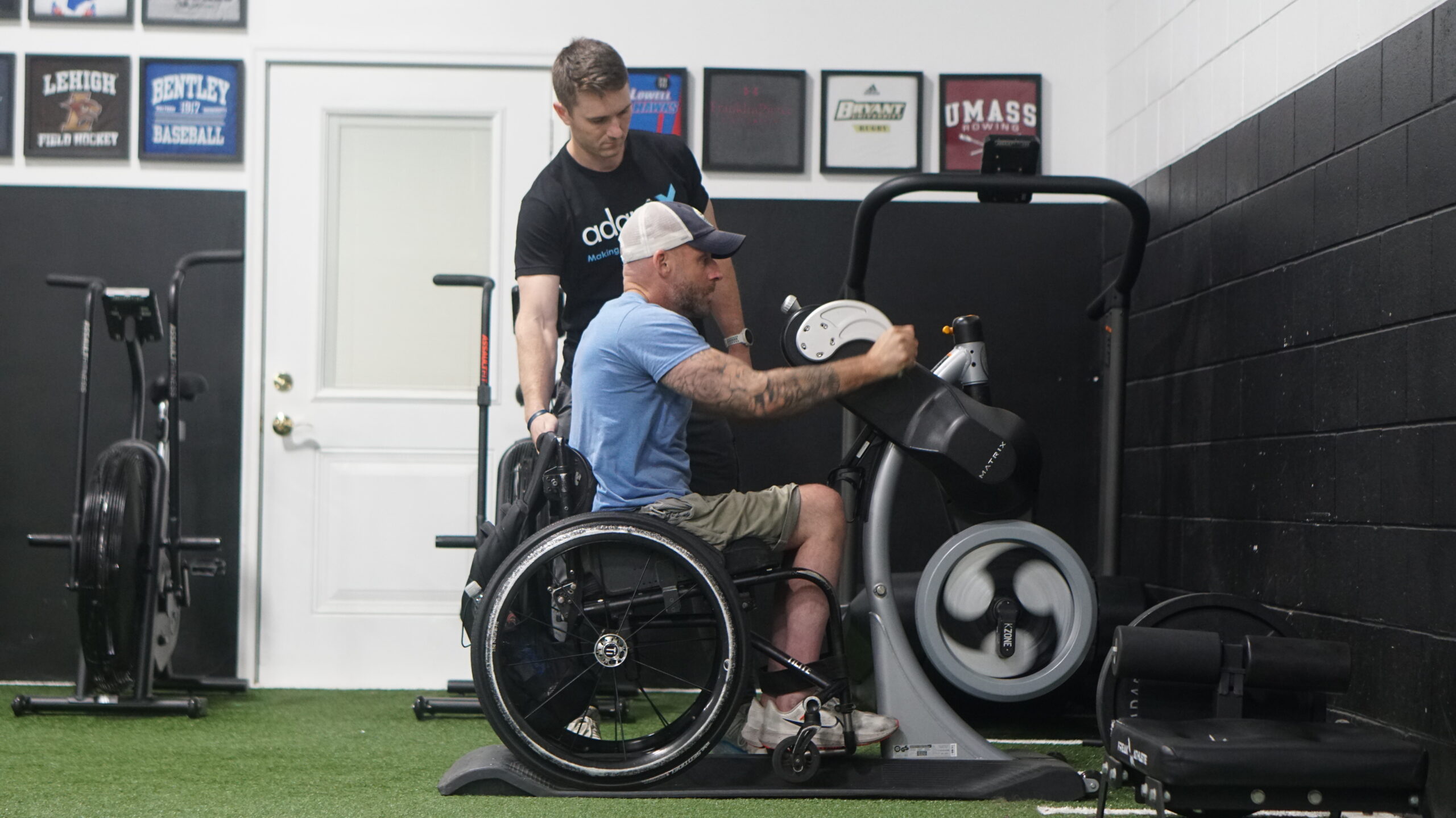 A man in a wheelchair uses an upper-body ergometer in a gym, gripping the hand pedals and focusing on his workout. He wears a baseball cap, a blue sleeveless shirt, and shorts, revealing tattooed arms. Behind him, a trainer in a black "adaptX" shirt stands attentively, providing support. The gym has a turf floor, black walls, and framed college sports banners. Various exercise machines and equipment are visible in the background.