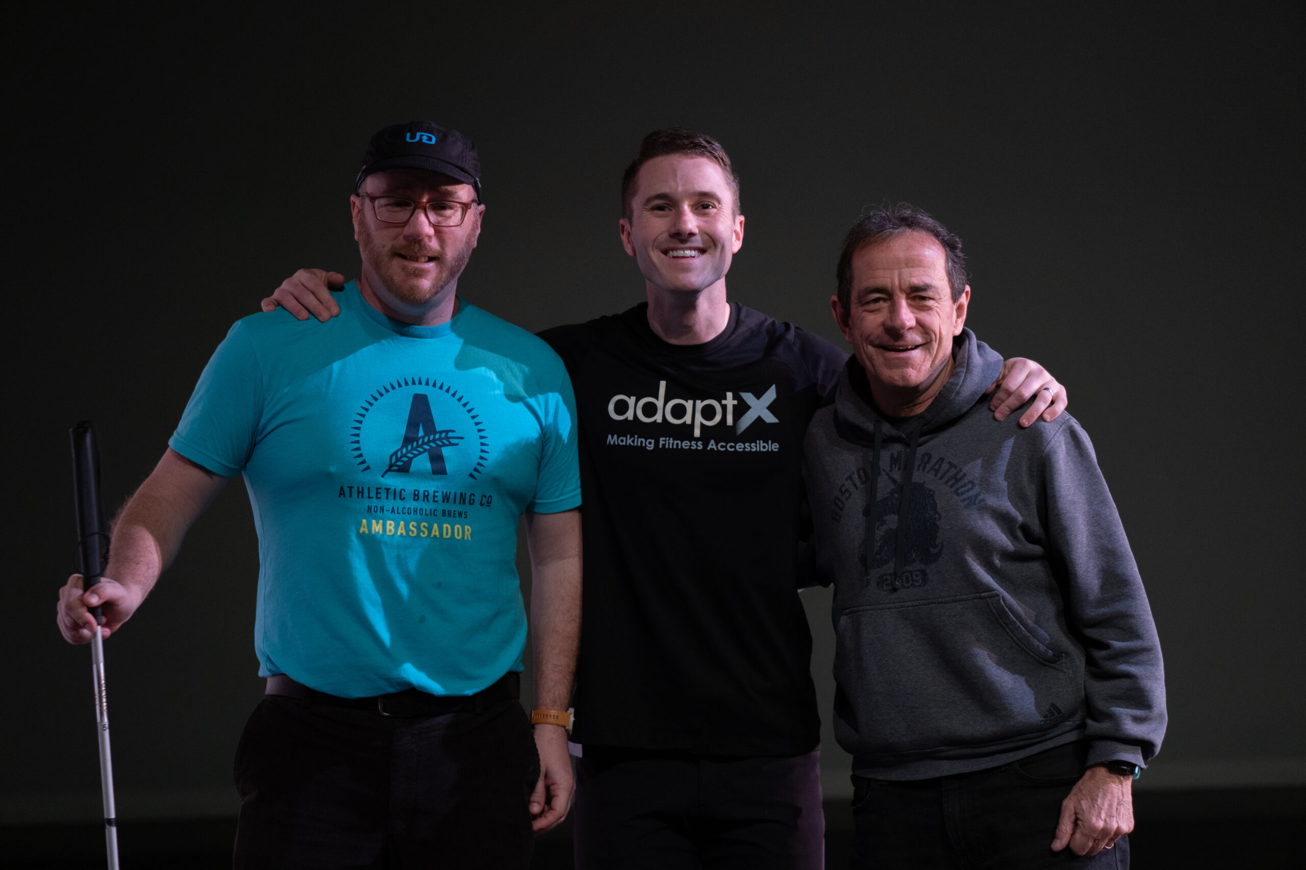 Three men stand close together, smiling at the camera against a dark background. The man on the left wears a blue "Athletic Brewing Co. Ambassador" T-shirt, a black cap, and glasses, holding a white cane. The man in the middle wears a black "AdaptX" T-shirt and has his arms around the shoulders of the other two. The man on the right wears a gray "Boston Marathon" hoodie and has his arm around the middle man's back. They appear to be in a dimly lit indoor setting.