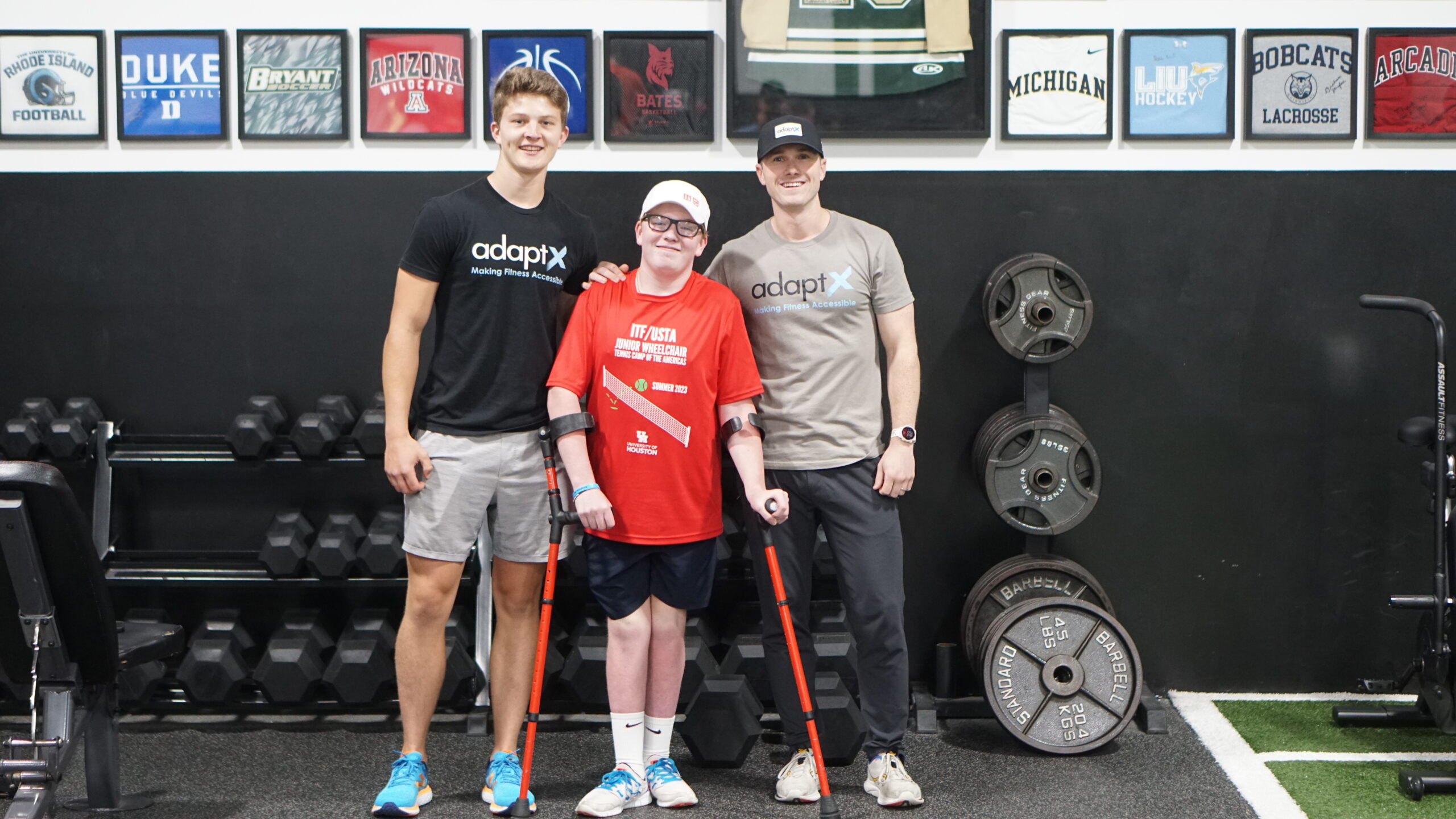 Three individuals pose together in a gym, smiling at the camera. The person in the center, wearing a red ITF/USTA Junior Wheelchair Tennis Tournament shirt and a white cap, stands with the support of red forearm crutches. The two individuals on either side wear AdaptX shirts, with one in black and the other in beige, both placing a supportive hand on the shoulders of the person in the center. Behind them, dumbbells, weight plates, and framed sports memorabilia are visible on the gym walls.