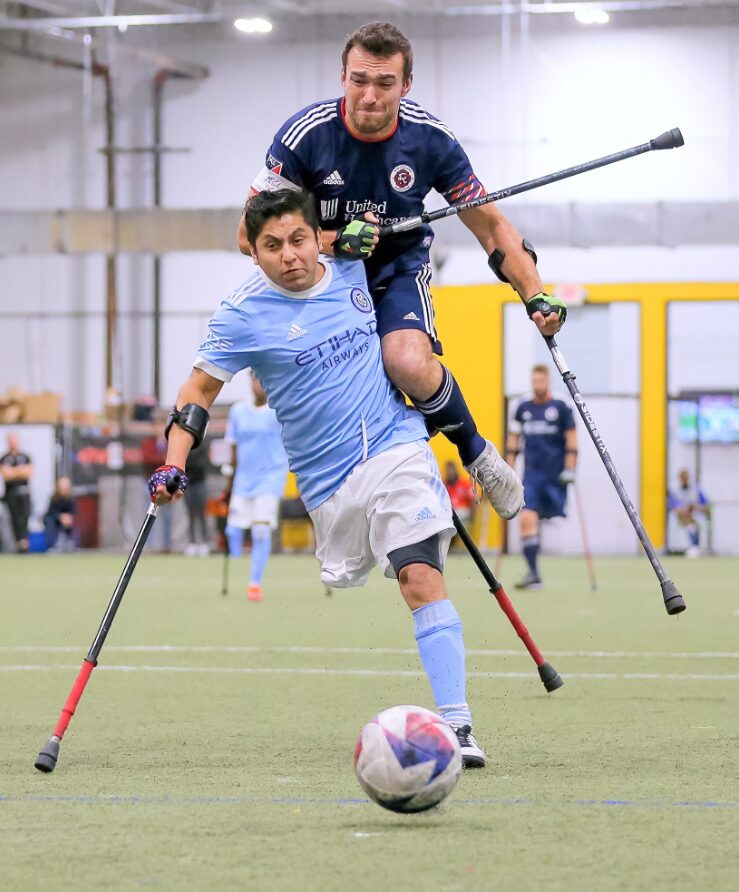 Two amputee soccer players compete for control of the ball during a match in an indoor sports facility. The player in the light blue "NYCFC" jersey moves forward using forearm crutches, focusing on the ball. The player in the navy blue "New England Revolution" jersey leaps onto his opponent’s back while also using crutches, showing intense determination. In the background, other players and spectators watch the fast-paced game. The artificial turf field and bright lighting highlight the competitive atmosphere.