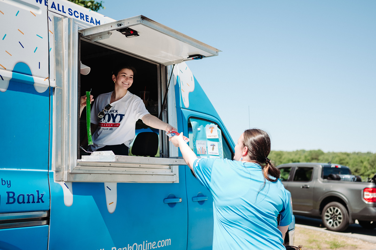 A smiling woman inside a bright blue ice cream truck hands a frozen treat to a customer wearing a matching blue shirt. The woman in the truck wears a white "Rick Hoyt Research Lab" t-shirt and holds another frozen treat in her other hand. The truck features ice cream graphics and text, with a menu displayed on the side. A pickup truck is parked in the background under a clear blue sky.