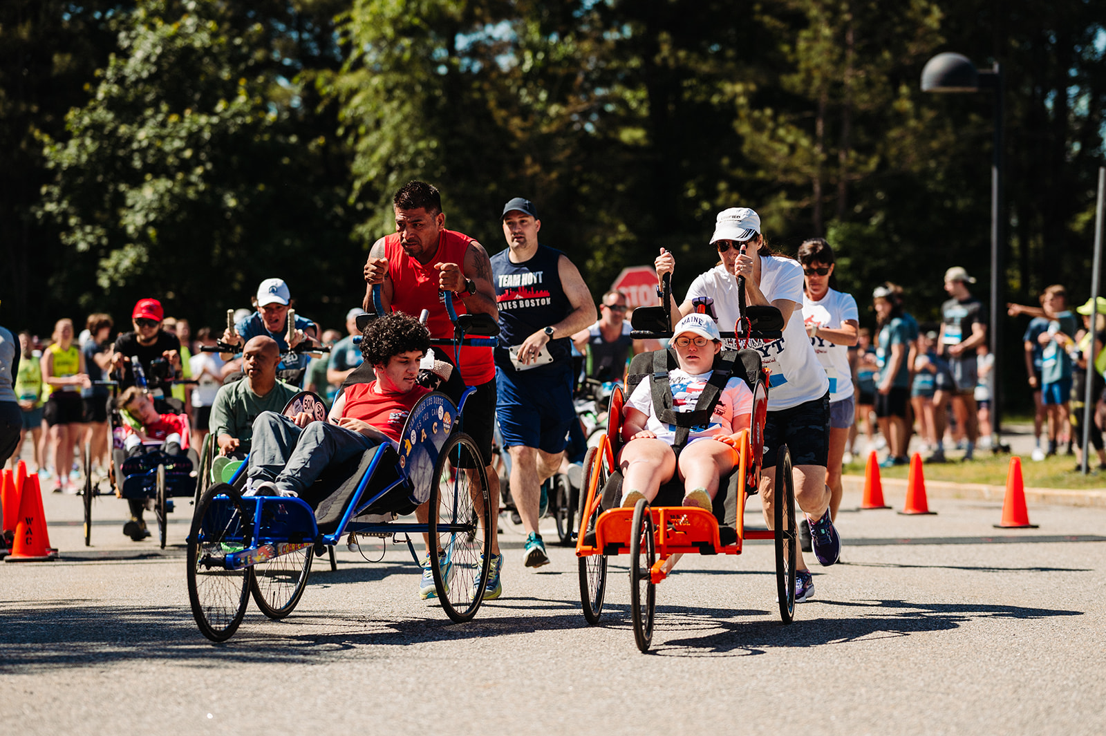 A group of athletes and volunteers participating in an inclusive race event. In the foreground, two push-assisted racing wheelchairs are being propelled by their running partners. One athlete in a red racing chair is wearing a white hat and sunglasses, while another in a blue racing chair is being pushed by a runner in a red tank top. The background features a crowd of spectators, additional racers, and volunteers, with orange cones marking the race route under a sunny sky.