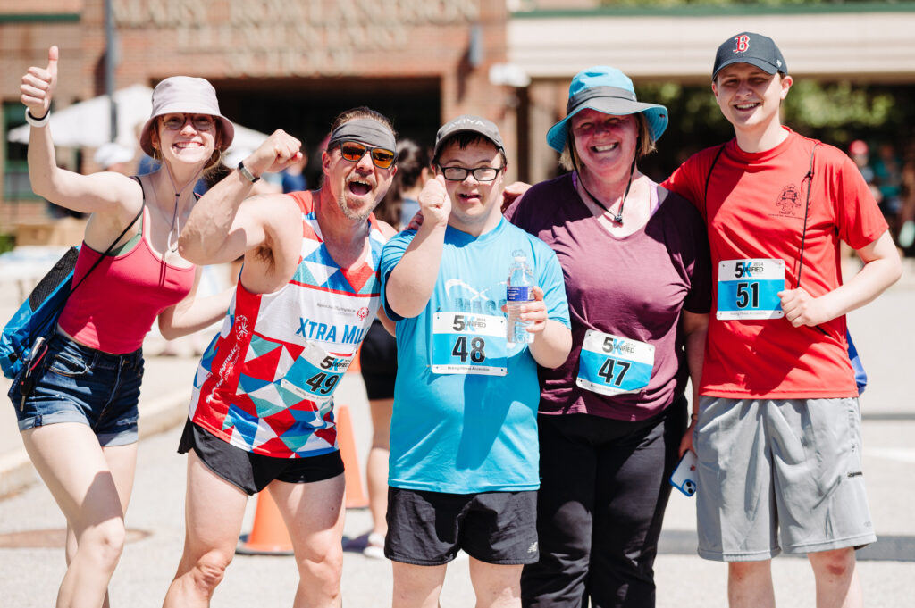 A group of five people pose together, smiling and cheering at an outdoor 5K race event. They are wearing race bibs numbered 47 to 51 and are dressed in athletic and casual attire. One person in the center, wearing a blue shirt with bib number 48, holds a water bottle. To their left, a man in a red, white, and blue running tank top flexes his arm enthusiastically. The others, including a woman in a sun hat and a young man in a red shirt and baseball cap, also smile and celebrate. The background features a sunny outdoor setting with race tents and a brick building.