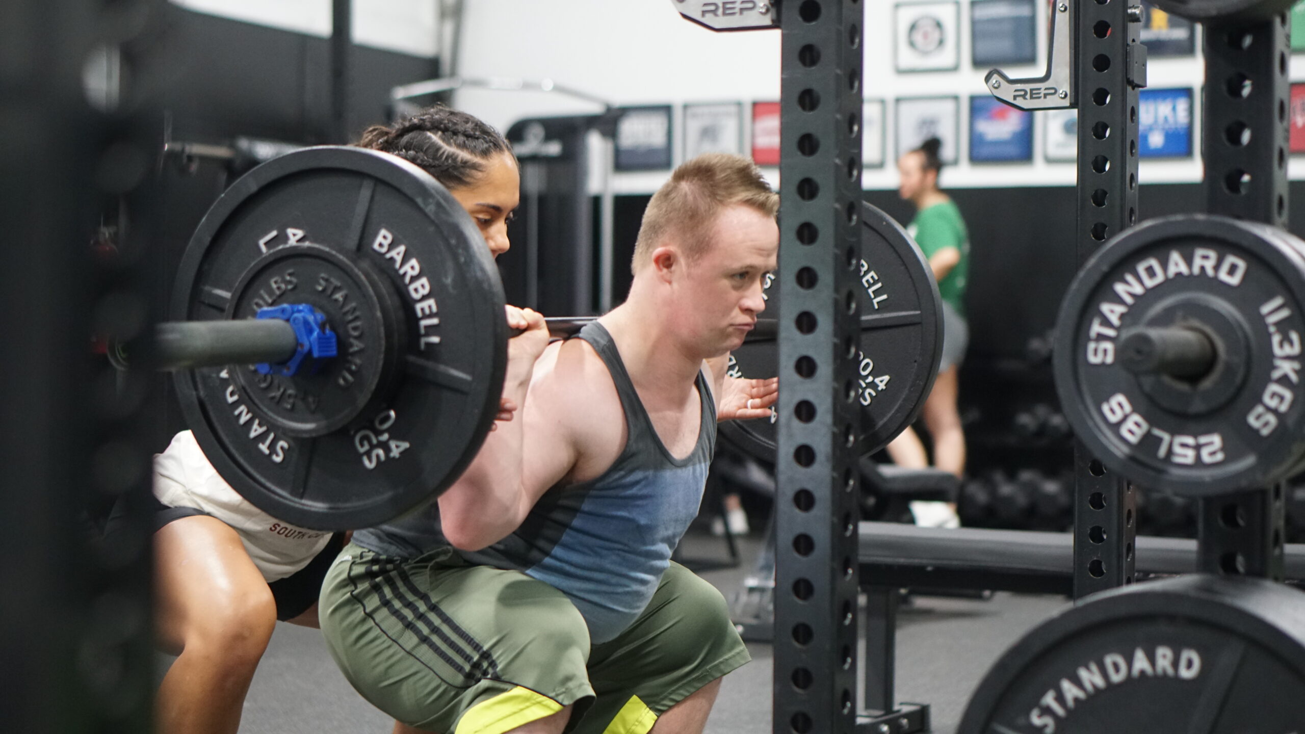 The image captures a young man with Down syndrome performing a barbell squat in a gym, with a coach spotting him closely. He is wearing a sleeveless workout top and green athletic pants with yellow knee sleeves. His expression is focused and determined as he lifts the weighted barbell. The coach, standing behind him, is providing support by keeping her hands near the bar. The gym is equipped with weightlifting racks, barbells, and various strength training equipment. In the background, other individuals are working out, and framed athletic memorabilia decorate the walls. The scene highlights inclusive fitness training and strength development.