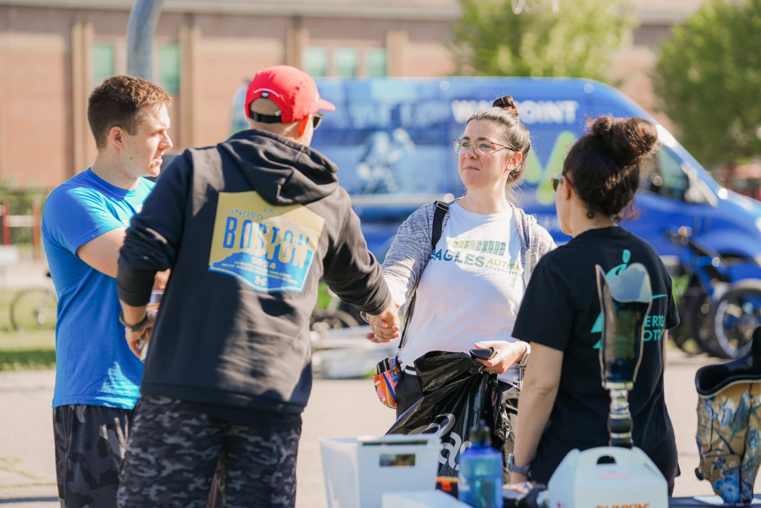 A man in a black hoodie with a 'Boston' logo and a red cap shakes hands with a woman wearing glasses and a white 'Eagles Autism Foundation' shirt. She carries a black bag and holds a phone in her other hand. Another woman in a black shirt with a green apple logo stands nearby, next to a table displaying a prosthetic leg, a water bottle, and other items. In the background, a man in a blue shirt gestures while speaking, and a blue van with a partially visible logo is parked near the event area.