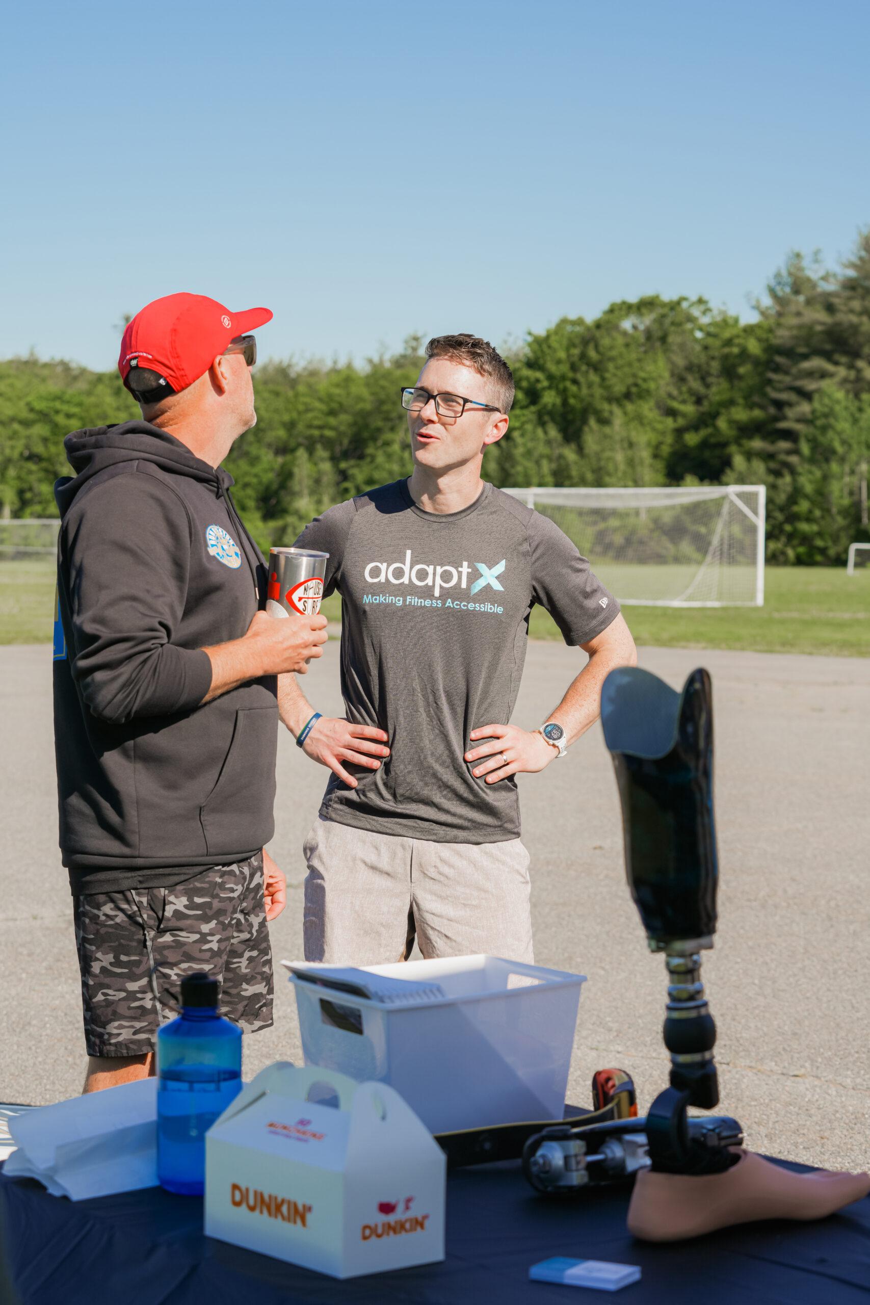 A man wearing a gray 'adaptX' shirt and glasses smiles while talking to another man in a black hoodie and red cap, who holds a can of soda. They stand near a table with a prosthetic leg, a Dunkin' box, a water bottle, and other items. Behind them, a soccer field and lush green trees are visible under a clear sky.