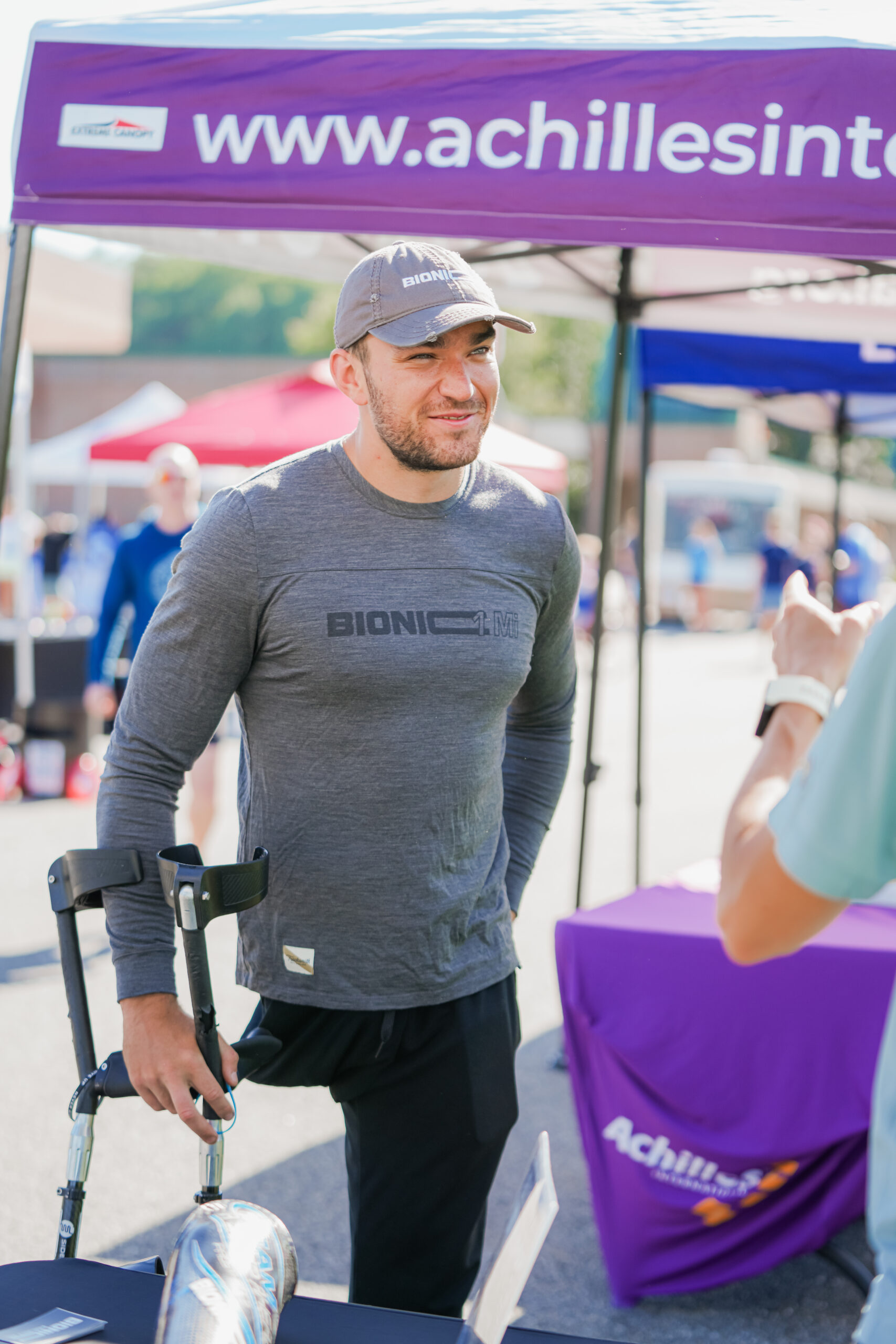 A man wearing a gray 'Bionic' long-sleeve shirt and cap smiles while standing under a tent with the 'Achilles International' website printed on it. He holds onto forearm crutches, and a prosthetic running blade is visible on a table nearby. Another person in a light blue shirt gestures while speaking to him. The background features other event attendees and booths at an outdoor gathering.