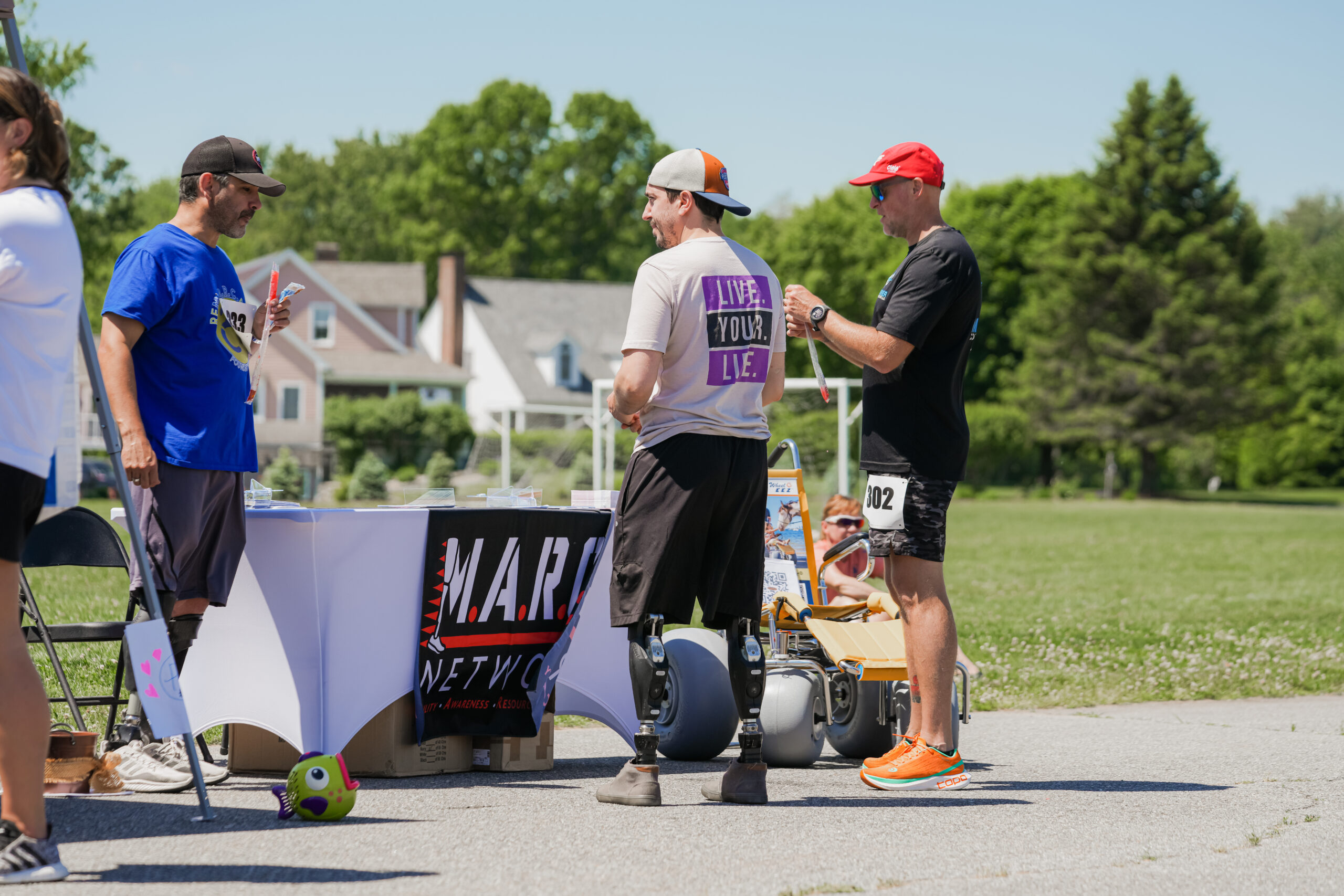 A group of people gathers around an outdoor event booth with a banner reading "M.A.R. NETWORK." In the foreground, a man wearing a white cap, a T-shirt that says "LIVE. YOUR. LIE." on the back, and athletic shorts stands with his back to the camera, revealing two prosthetic legs. He is engaged in conversation with another man in a black T-shirt, race bib number 302, and bright orange running shoes, who is holding a freeze pop. To the left, a man in a blue shirt and black cap is eating a snack while standing near the booth. Behind them, a woman sits in a yellow all-terrain wheelchair. The background features green grass, trees, and houses under a bright blue sky.