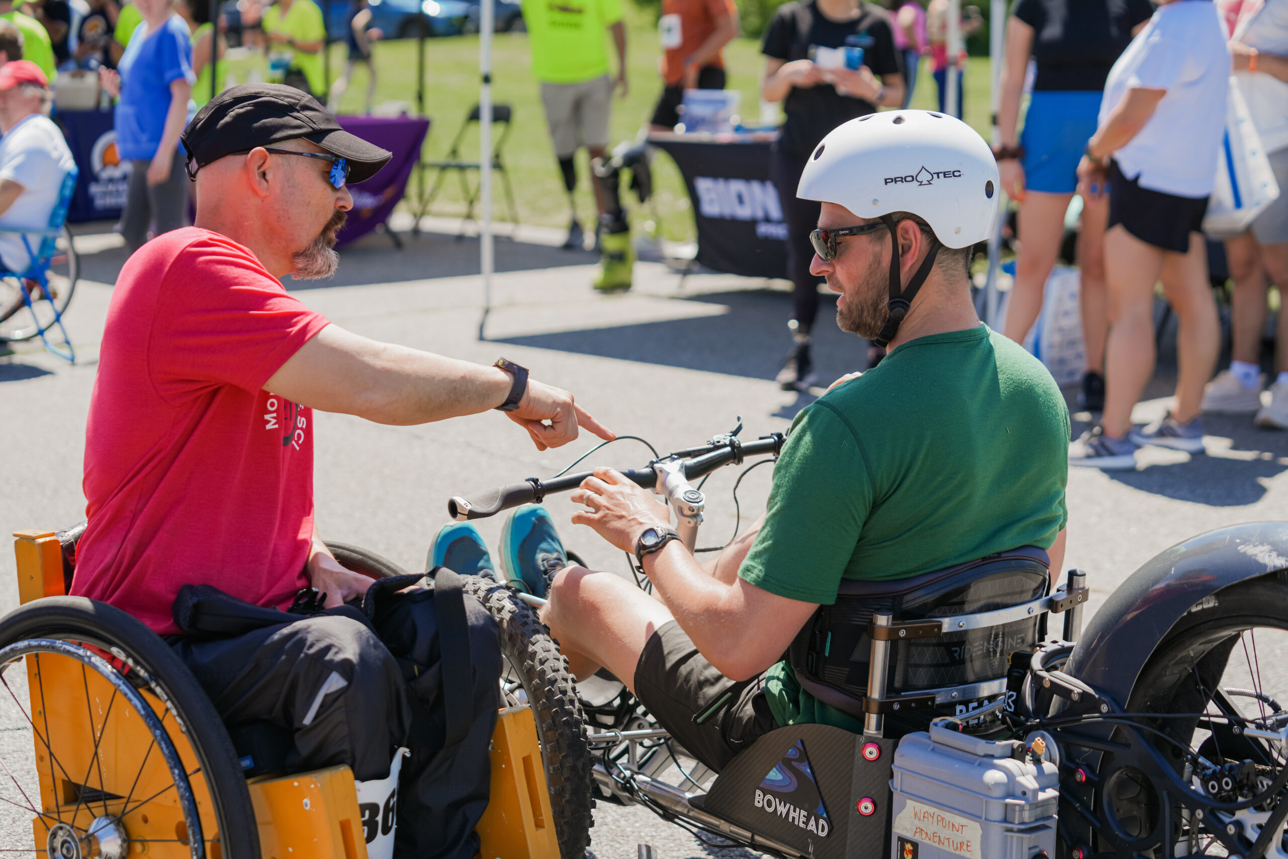 Two men are engaged in conversation at an outdoor event, both using adaptive mobility equipment. The man on the left, wearing a red t-shirt, black cap, and sunglasses, is seated in a yellow wheelchair and is gesturing while speaking. The man on the right, wearing a green t-shirt, black shorts, and a white helmet labeled "Pro-Tec," is seated on a Bowhead adaptive bike with large wheels and a battery pack labeled "Waypoint Adventure." He listens with his hands resting on the handlebars. The background features a crowd of people, event tents, and banners.