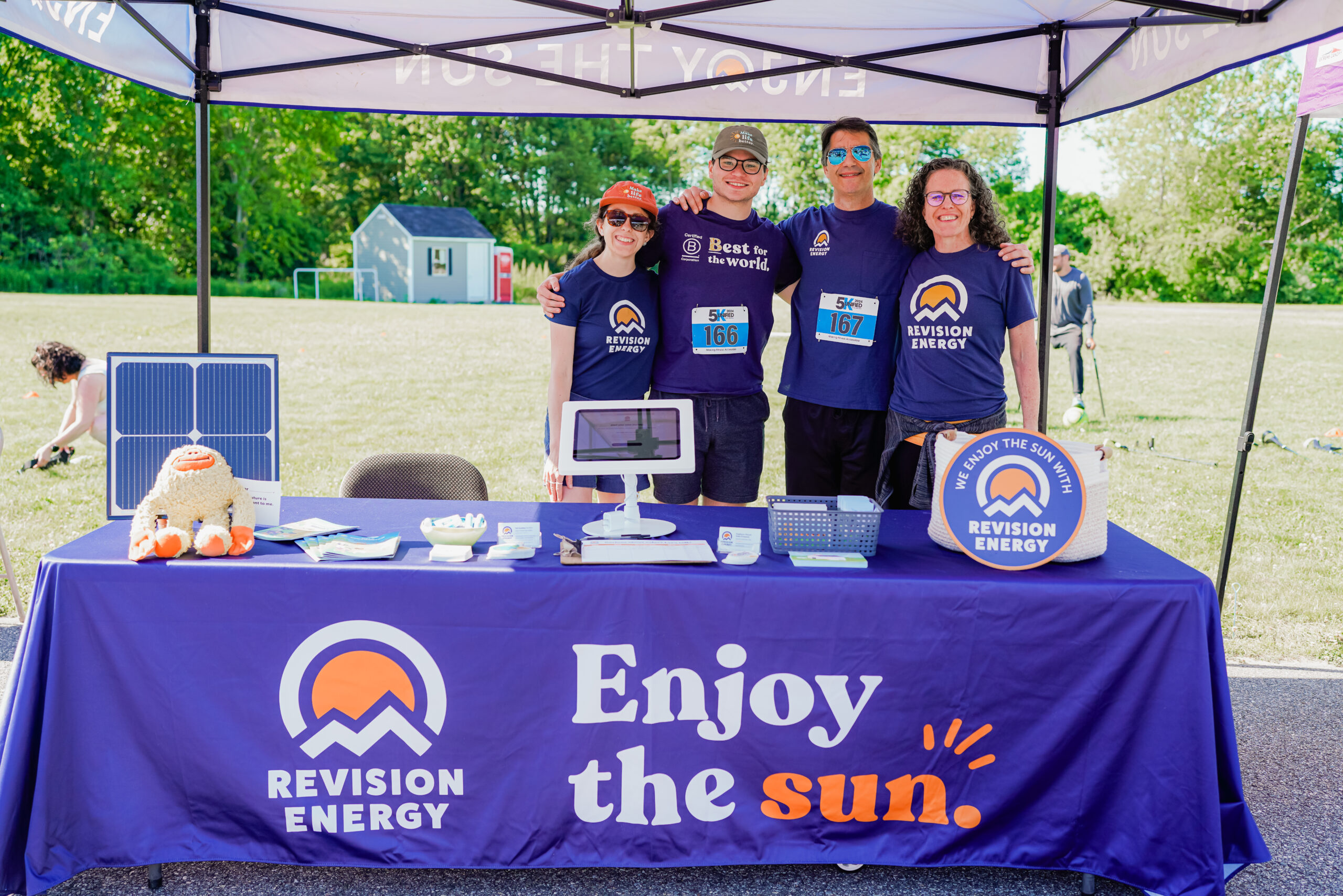 A group of four people stands together, smiling under a tent promoting Revision Energy. They are wearing matching navy blue shirts with the company logo, and two of them have race bibs. The booth setup includes a table covered with a blue cloth displaying the words "Enjoy the sun." Various promotional materials, a small solar panel, and a plush mascot are arranged on the table. The background features a grassy field, a small building, and event attendees.