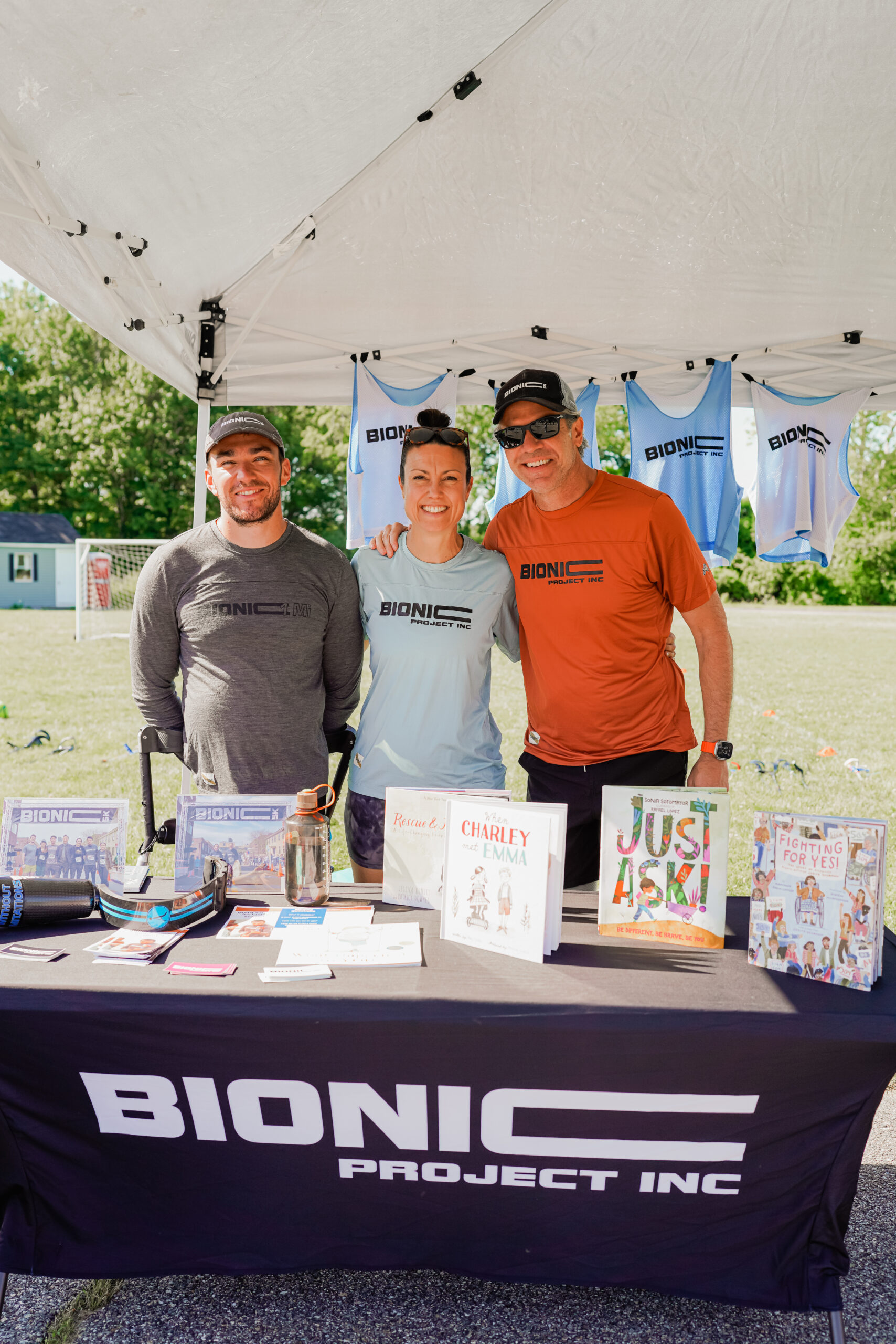 Three people stand together under a white event tent, smiling at the camera. They are representing BIONIC Project Inc., as indicated by the branded tablecloth and shirts they are wearing. The table in front of them displays books, pamphlets, and a prosthetic running blade. Behind them, blue BIONIC-branded tank tops hang from the tent. The outdoor setting features a grassy field with other event booths and attendees visible in the background.