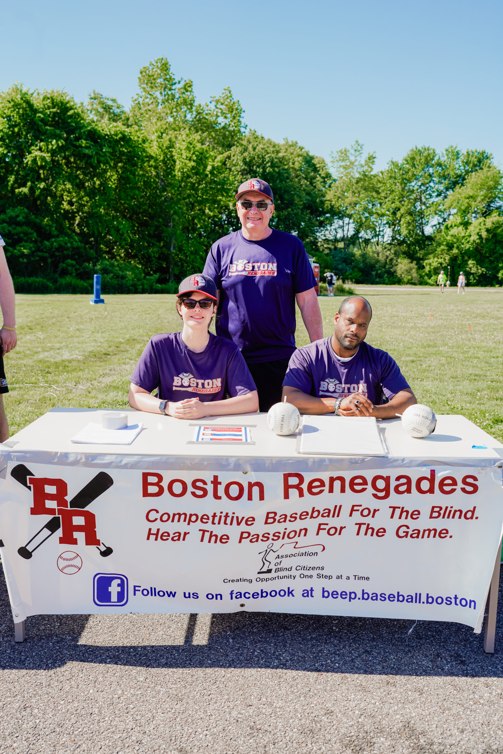 Three members of the Boston Renegades, a competitive baseball team for the blind, pose at an outdoor event on a sunny day. Two individuals are seated at a table covered with a white banner displaying the team's name, logo, and slogan: "Hear the Passion For The Game." The seated individuals wear matching purple "Boston Renegades" shirts, with one wearing sunglasses and a baseball cap, while the other rests their hands on the table near two baseballs. A third team member, also wearing a team shirt and sunglasses, stands behind them smiling. The table has promotional materials, and a grassy field with trees and event attendees is visible in the background.