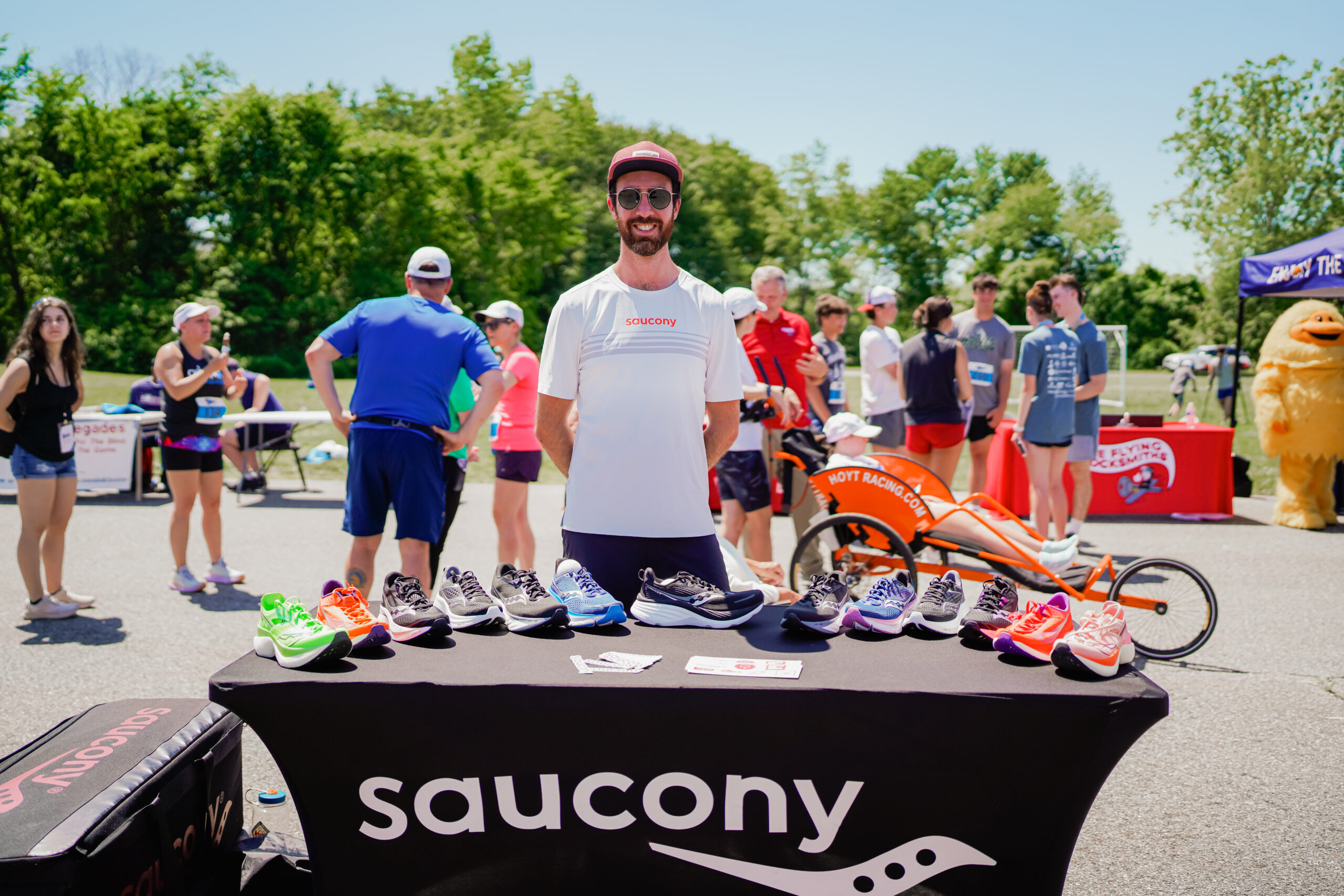 "A smiling man wearing sunglasses, a red cap, and a white 'Saucony' shirt stands behind a table displaying an array of colorful running shoes at an outdoor event. The table has a black cloth with the 'Saucony' logo. In the background, people are gathered around, some in athletic gear, while an orange racing wheelchair with 'Hoyt Racing' branding is visible. A mascot in a yellow costume stands near a tent labeled 'Flying' with a red banner.