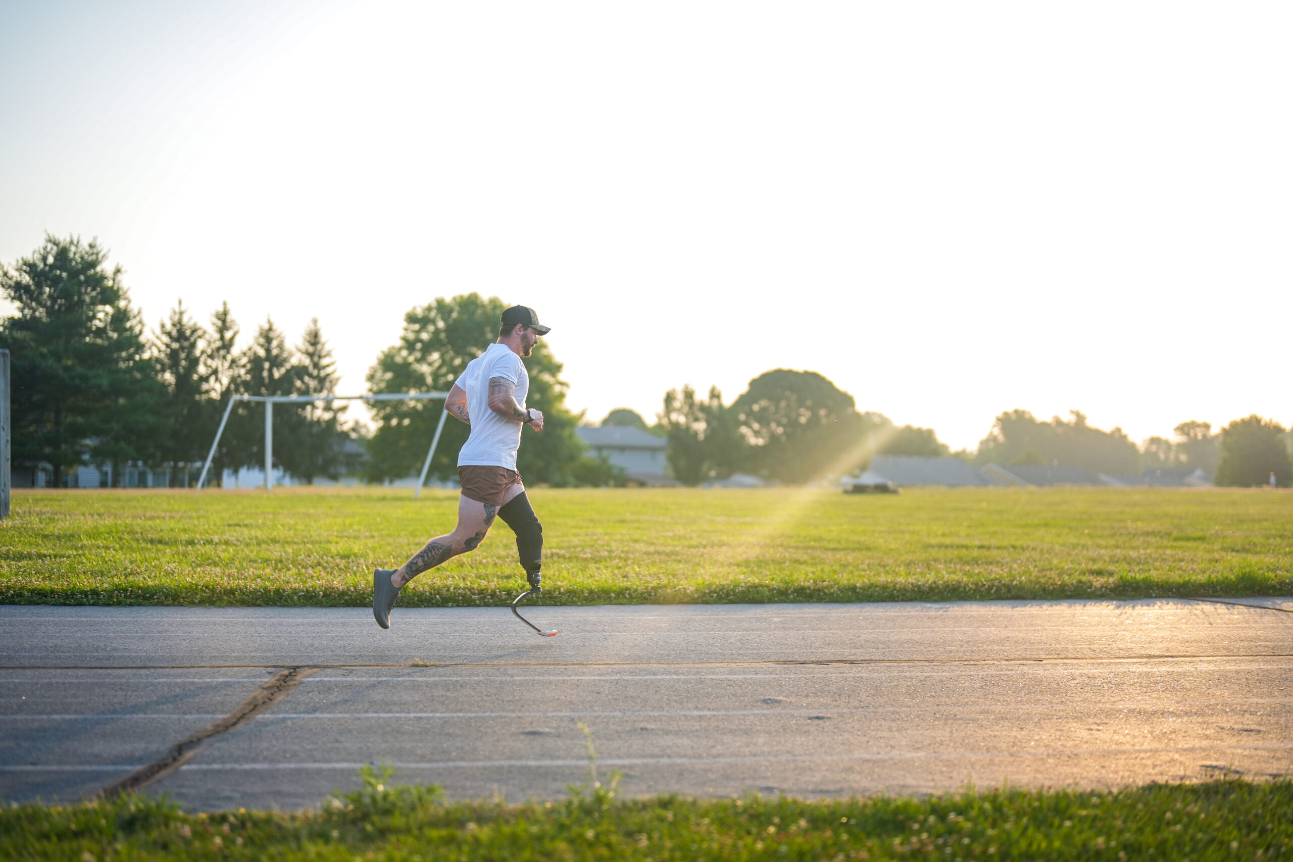 A male runner with a prosthetic blade on his right leg is mid-stride on a paved path, illuminated by the golden light of the setting sun. He wears a white t-shirt, brown shorts, a black cap, and running shoes, with his arms pumping in rhythm. The background features an open grassy field, a soccer goal, and distant trees, with warm sunlight streaming through the scene.