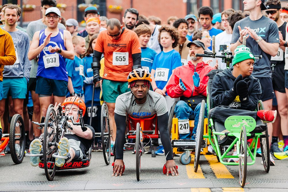 A diverse group of athletes, including wheelchair racers and handcyclists, lines up at the starting line of a race, with a large crowd of runners and spectators behind them. In the foreground, three adaptive athletes are focused and ready: one using a handcycle, another in a racing wheelchair wearing an orange helmet, and a third in a green wheelchair wearing a beanie and gloves. The background features runners in colorful athletic gear, many wearing race bibs, some clapping or cheering. The scene is energetic and inclusive, with a mix of athletes of different abilities preparing for the race.