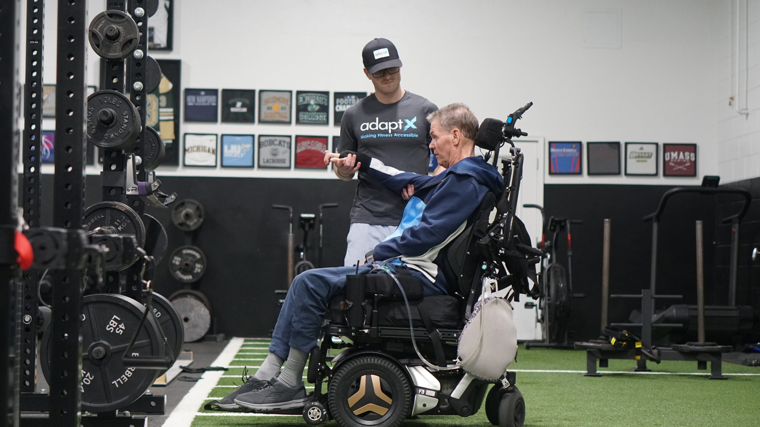 The image shows a personal trainer assisting an older man in a power wheelchair during a workout session at a gym. The trainer, wearing a black "adaptX" cap and t-shirt with the tagline "Making Fitness Accessible," is attentively guiding the man through an exercise using a resistance band. The older man is dressed in a navy blue tracksuit and sneakers, actively engaging in the movement. The gym is equipped with weight racks, barbells, and other fitness equipment, with a turf area for adaptive training. The walls are decorated with framed athletic memorabilia, creating an inclusive and motivational training environment.