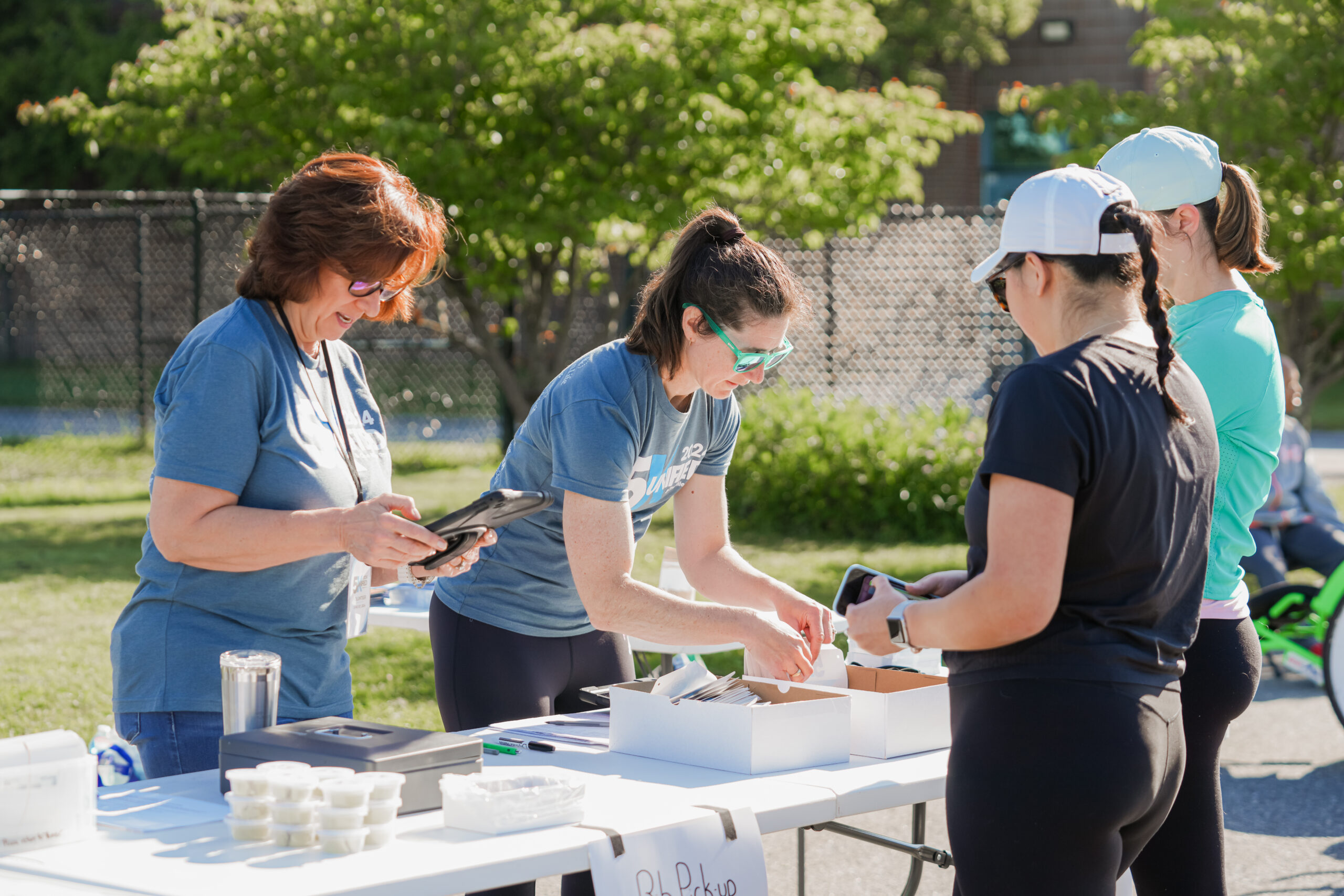 The image captures an outdoor event registration or check-in station where volunteers and participants are interacting. Two volunteers in blue event t-shirts are behind a white table, assisting attendees. One volunteer, wearing sunglasses, is using a tablet, while the other is distributing race bibs or materials from a box. On the opposite side of the table, two women dressed in athletic gear, including hats and sunglasses, are receiving event materials. The table has labeled boxes, small cups, and other supplies, suggesting a race or fitness event check-in. The background features a fenced grassy area with trees and a building, indicating a park or school setting. The bright sunlight and shadows suggest a warm, sunny day, and the scene conveys an atmosphere of community engagement and preparation for a fitness event. The image captures an outdoor event registration or check-in station where volunteers and participants are interacting. Two volunteers in blue event t-shirts are behind a white table, assisting attendees. One volunteer, wearing sunglasses, is using a tablet, while the other is distributing race bibs or materials from a box. On the opposite side of the table, two women dressed in athletic gear, including hats and sunglasses, are receiving event materials. The table has labeled boxes, small cups, and other supplies, suggesting a race or fitness event check-in. The background features a fenced grassy area with trees and a building, indicating a park or school setting. The bright sunlight and shadows suggest a warm, sunny day, and the scene conveys an atmosphere of community engagement and preparation for a fitness event