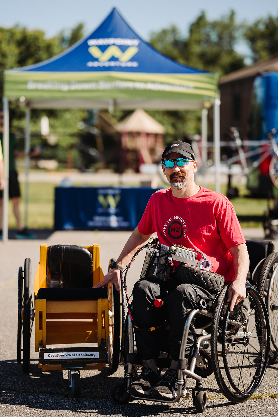 The image features a man in a wheelchair at an outdoor event, smiling and wearing a red "Move to Improve" t-shirt, black pants, sunglasses, and a black cap. He is sitting next to a custom-built adaptive wheelchair labeled "OpenSourceWheelchairs.org," which has a yellow seat and black frame. In the background, there is a blue and green tent with a "Wheelchair Sports" logo, along with a tagline that reads, "Breaking Down Barriers to Adaptive Sports." Other adaptive equipment, such as bicycles and additional wheelchairs, can be seen nearby. The bright sunlight and clear sky indicate a warm, sunny day, and the setting suggests an inclusive sports or mobility-focused event promoting accessibility and adaptive athletics.