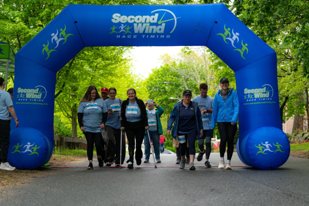 A diverse group of participants, including individuals using canes, walk together under a bright blue Second Wind Race Timing arch at the start of the adaptX Unified 5K.