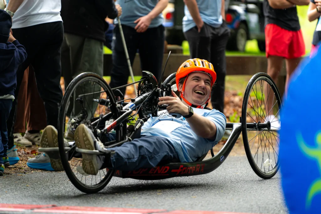A smiling participant wearing an orange helmet rides a handcycle past the finish line at the adaptX Unified 5K, surrounded by cheering spectators.