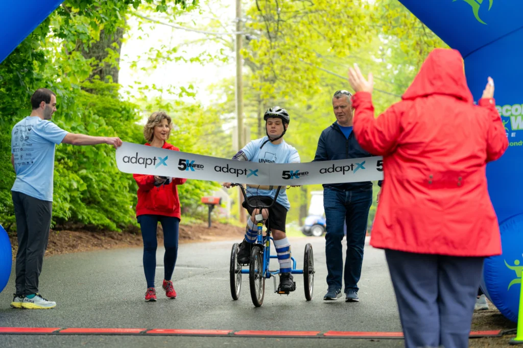 A young athlete using a handcycle crosses the finish line at the adaptX Unified 5K, flanked by two supporters and cheered on by volunteers under a large blue inflatable arch.