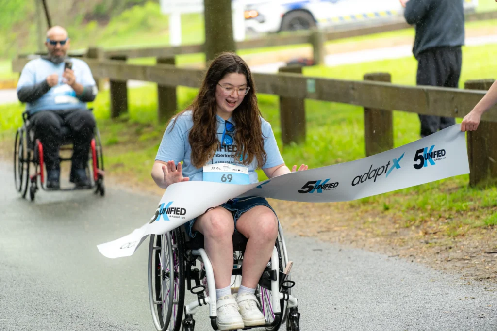 A young woman in a wheelchair wearing a race bib and “Unified 5K” shirt smiles as she crosses the finish line, breaking through the adaptX banner, with another participant behind her.