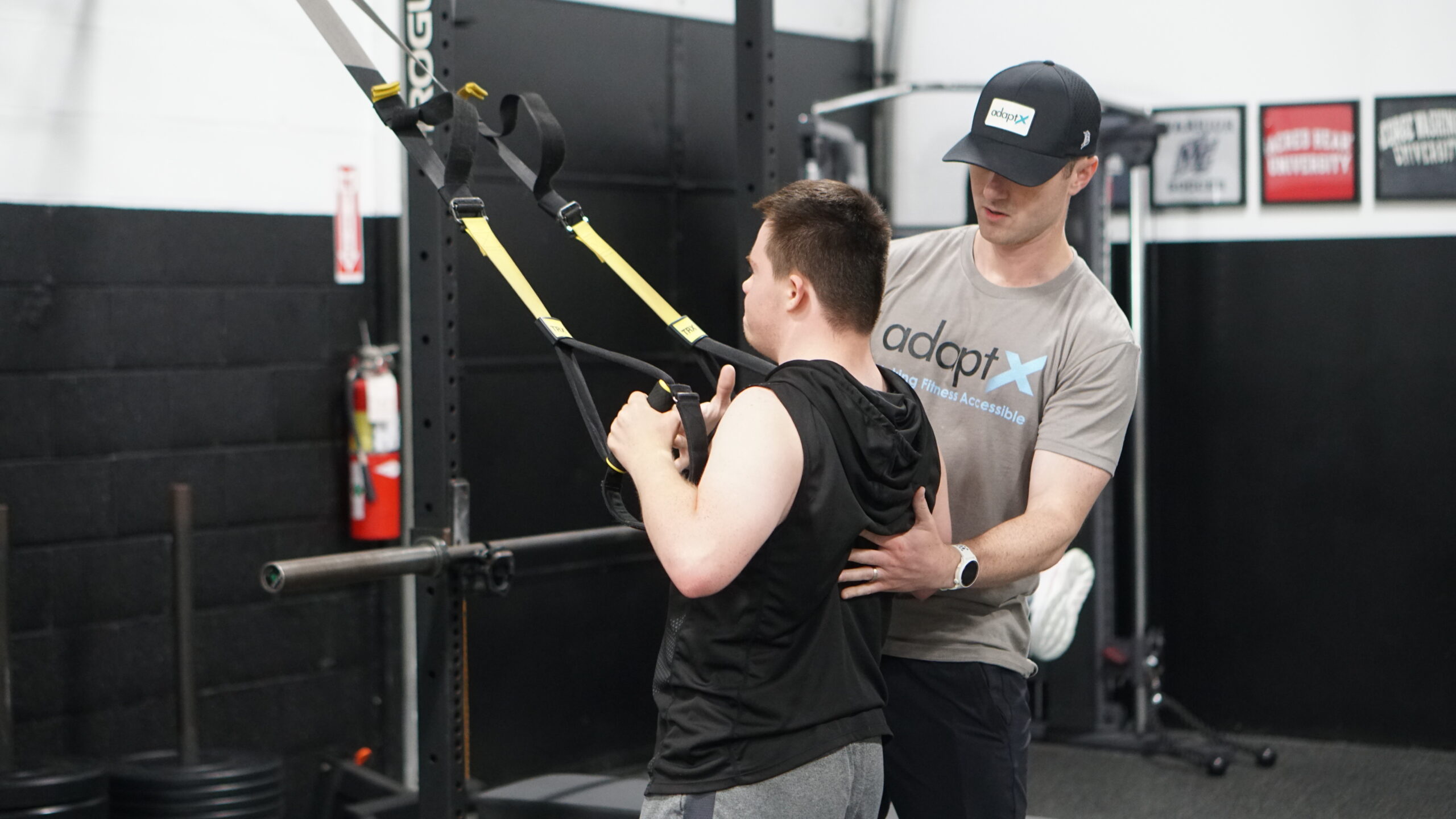 The image shows a fitness trainer assisting a young man with a TRX suspension training exercise in a gym. The trainer, wearing a gray "adaptX" t-shirt and a matching cap, provides support by placing a hand on the young man's back while he grips the TRX straps. The gym has a black-and-white color scheme, with equipment such as barbells, weight plates, and a squat rack visible in the background. There are framed posters on the wall, and a fire extinguisher is mounted nearby. The scene emphasizes inclusive fitness training and support for adaptive athletes.