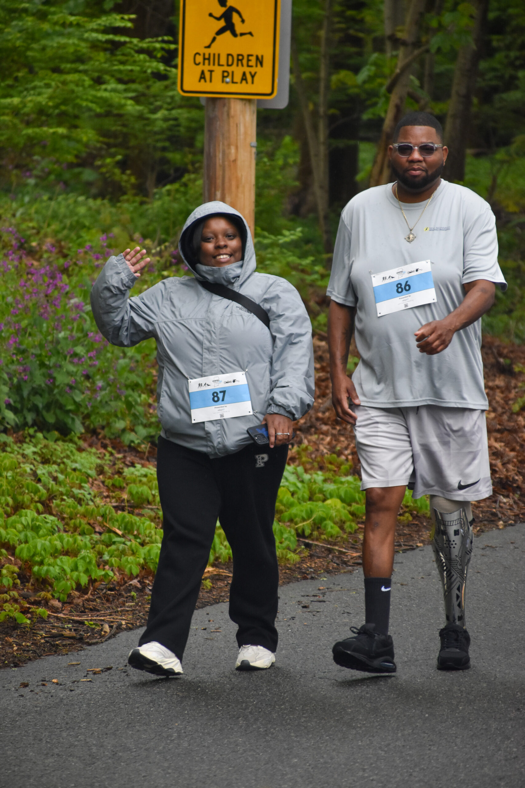 Two race participants walk along a wooded road wearing bib numbers 87 and 86; one waves and smiles, while the other, who has a prosthetic leg, walks beside her. A “Children at Play” sign is visible in the background.