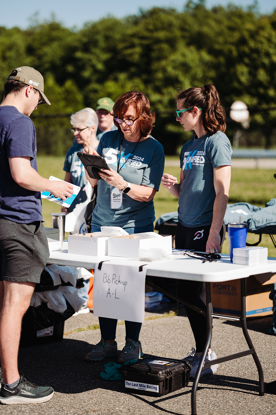 The image captures a race bib pickup station at an outdoor event. Two volunteers in matching blue "2024 Unified 5K" t-shirts are assisting a participant, who is dressed in athletic attire and wearing a cap. One volunteer is using a tablet, likely to check in participants, while the other is handing out race materials. A sign reading "Bib Pickup A-L" is attached to the table, which is covered with boxes, papers, and supplies. Behind the volunteers, other event staff and attendees are visible, some wearing hats and sunglasses. A black case labeled "The Last Mile Racing" is on the ground, possibly containing timing equipment. The setting is a sunny outdoor space, possibly a park or race venue, with a fenced grassy area and trees in the background. The scene highlights the organized and welcoming atmosphere of a community fitness event.