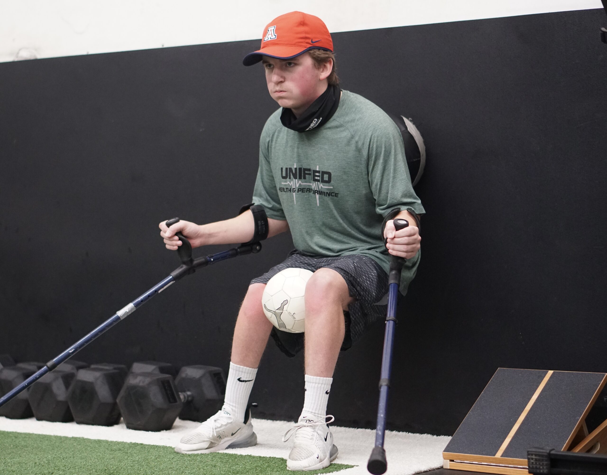 The image captures a young man performing a wall sit exercise while balancing a soccer ball between his knees. He is wearing a green "Unified" athletic performance t-shirt, gray shorts, white socks, and white sneakers. He also has on a red University of Arizona cap and a black neck gaiter. He is using forearm crutches, which are positioned at his sides for support. His expression is focused and determined. The setting is a gym with a black wall, a row of dumbbells, and adaptive training equipment visible in the background. The presence of specialized equipment suggests an inclusive fitness environment designed to support adaptive athletes.