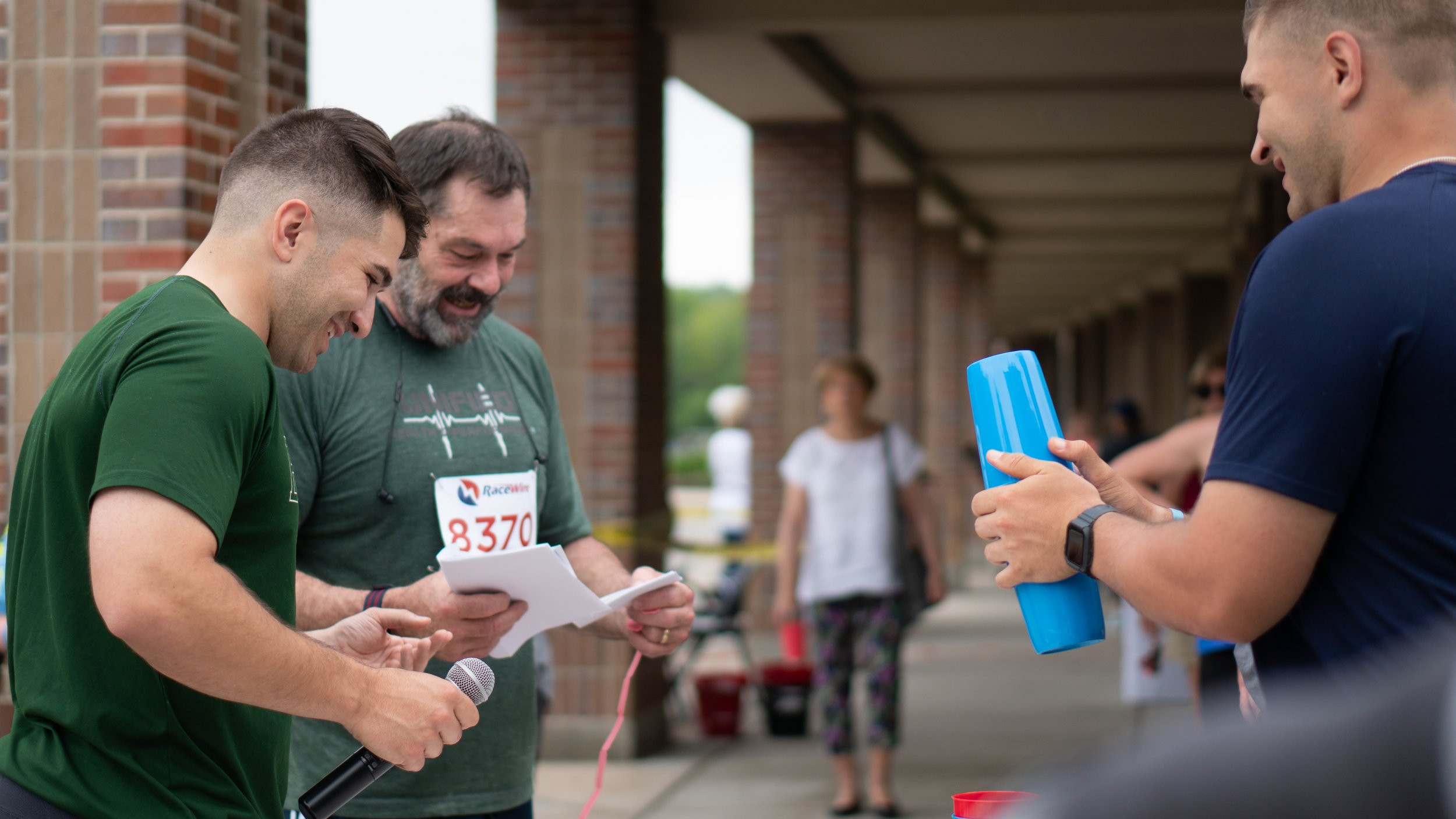 The image captures a joyful awards moment at an outdoor event, where a man in a green race t-shirt and bib number "8370" is receiving a prize. He is holding a paper and red ribbon, looking at them with a big smile. Next to him, another man in a dark green shirt holds a microphone, laughing and seemingly announcing the award. A third individual, wearing a navy blue shirt and smartwatch, is handing over a blue cylindrical prize to the race participant. The background features a covered walkway with brick pillars, and a few people walking or standing, dressed in casual summer attire. The setting suggests a community race or fitness event celebrating achievements with an engaging and positive atmosphere.