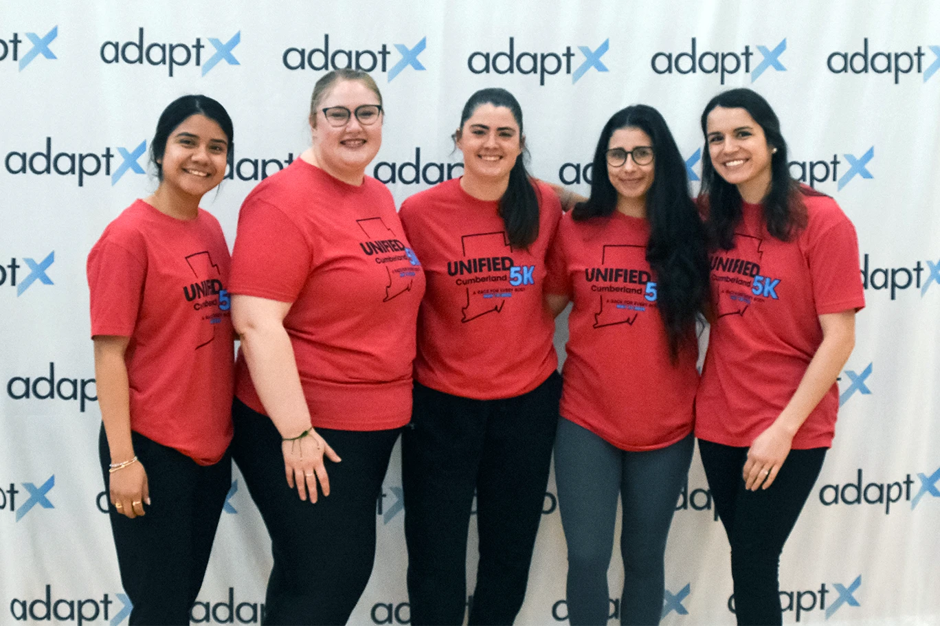 Five women in matching red “Unified 5K” shirts stand together and smile in front of an adaptX banner at an event.