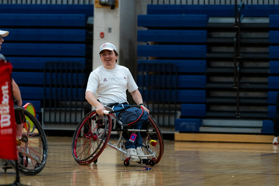 A smiling athlete in a sport wheelchair rolls across a gym floor, wearing a white shirt and cap, with bleachers and gym equipment in the background.