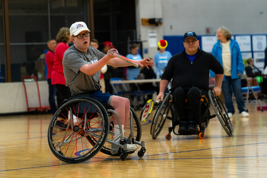 A young wheelchair athlete prepares to hit a ball with a paddle during an indoor adaptive sports event, as another participant and spectators look on in the gym.
