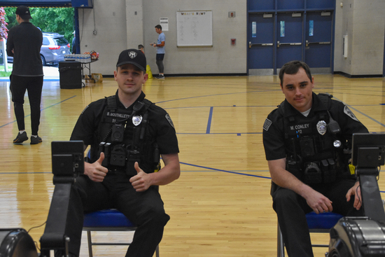 Two uniformed police officers sit side by side on rowing machines in a gymnasium; one gives a double thumbs-up while the other smiles slightly.
