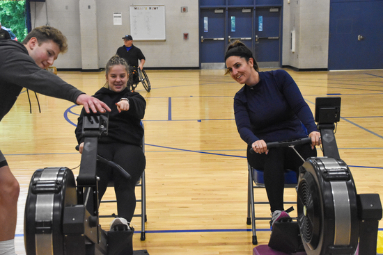 Two women on rowing machines in a gym smile as a young man shows them something on the display; a person in a wheelchair moves in the background.
