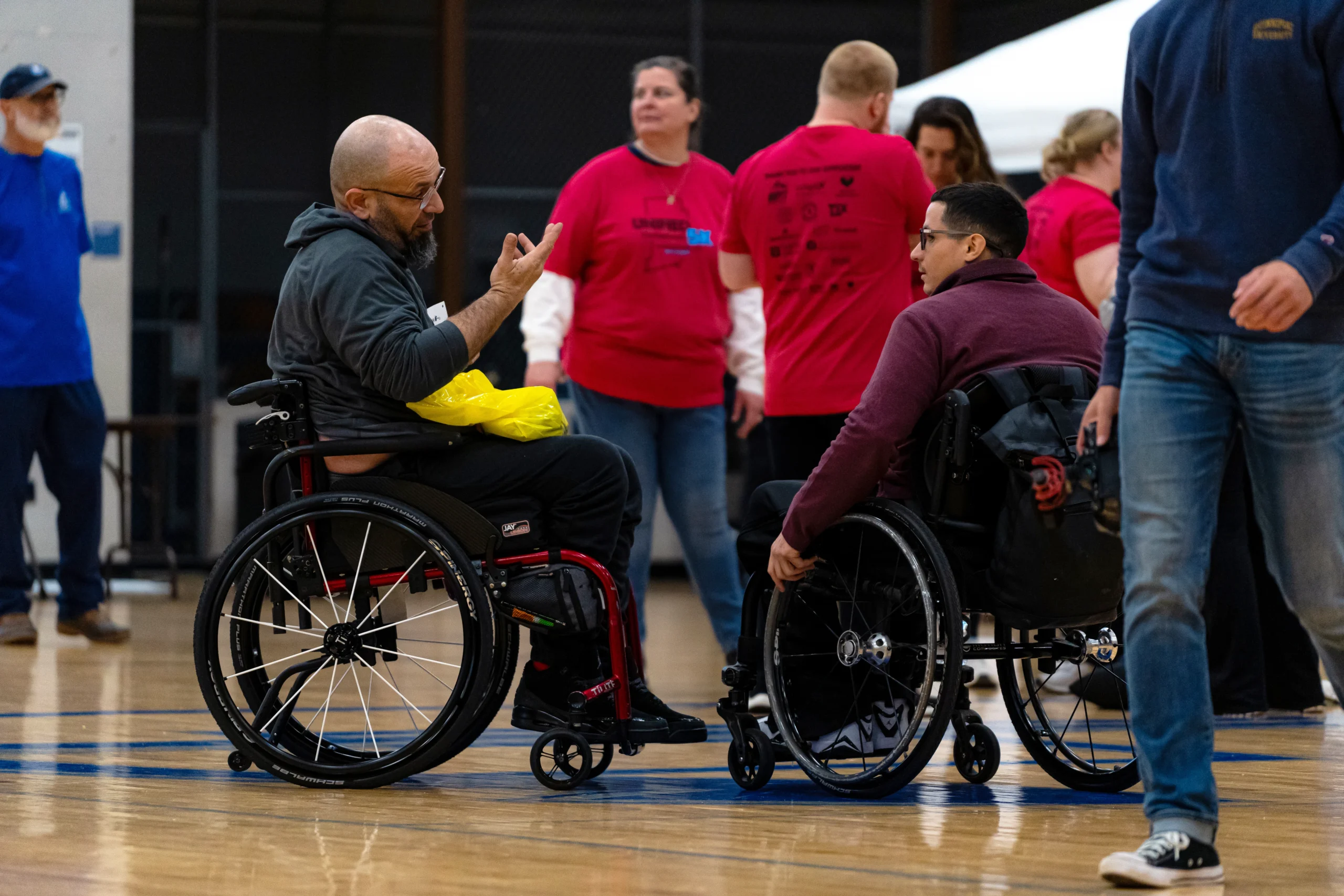 Two men using wheelchairs talk on a gym floor during an indoor event, with participants in red “Unified 5K” shirts and others moving around in the background.