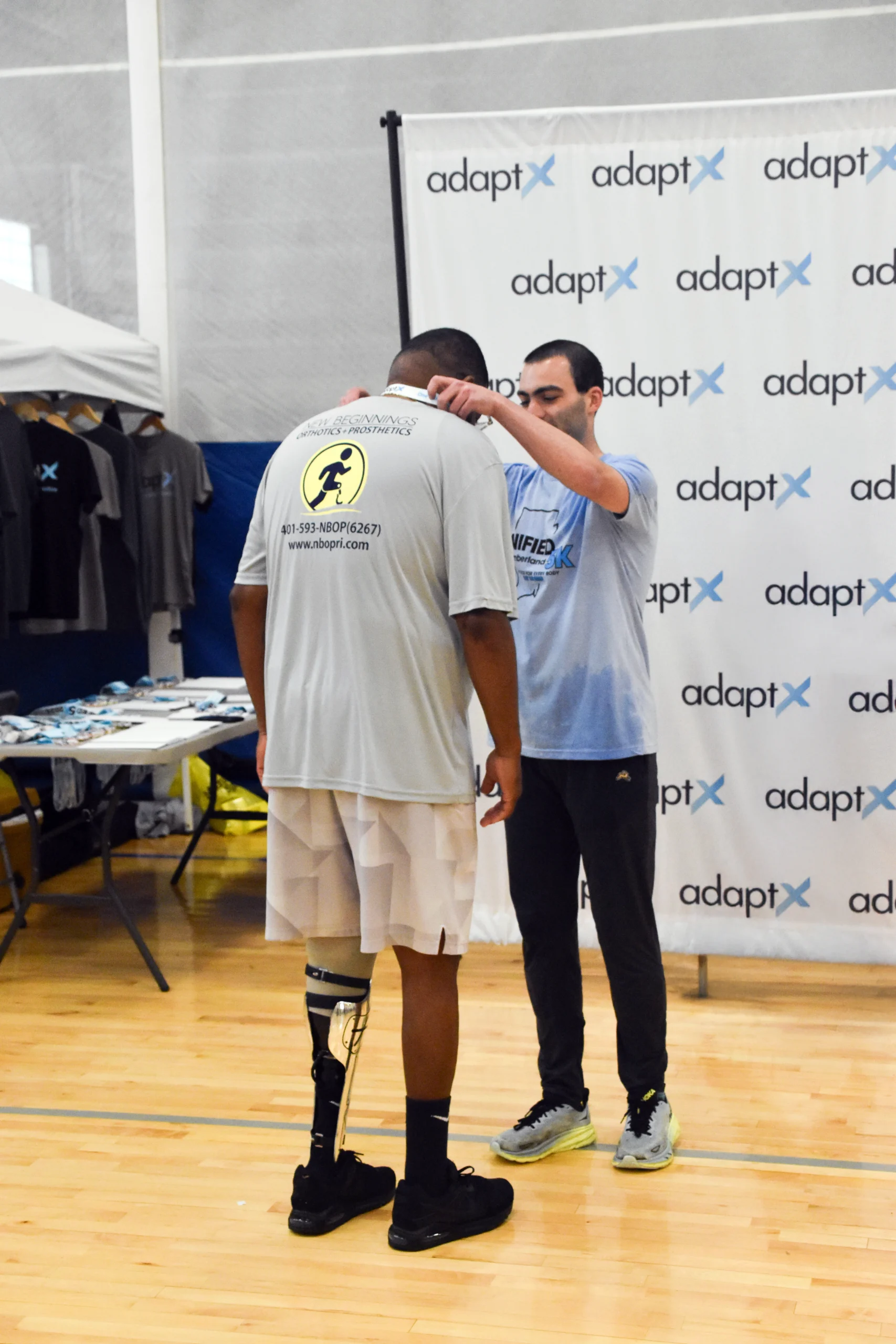 A man places a medal around the neck of a race participant wearing a prosthetic leg, in front of an adaptX backdrop at an indoor event.