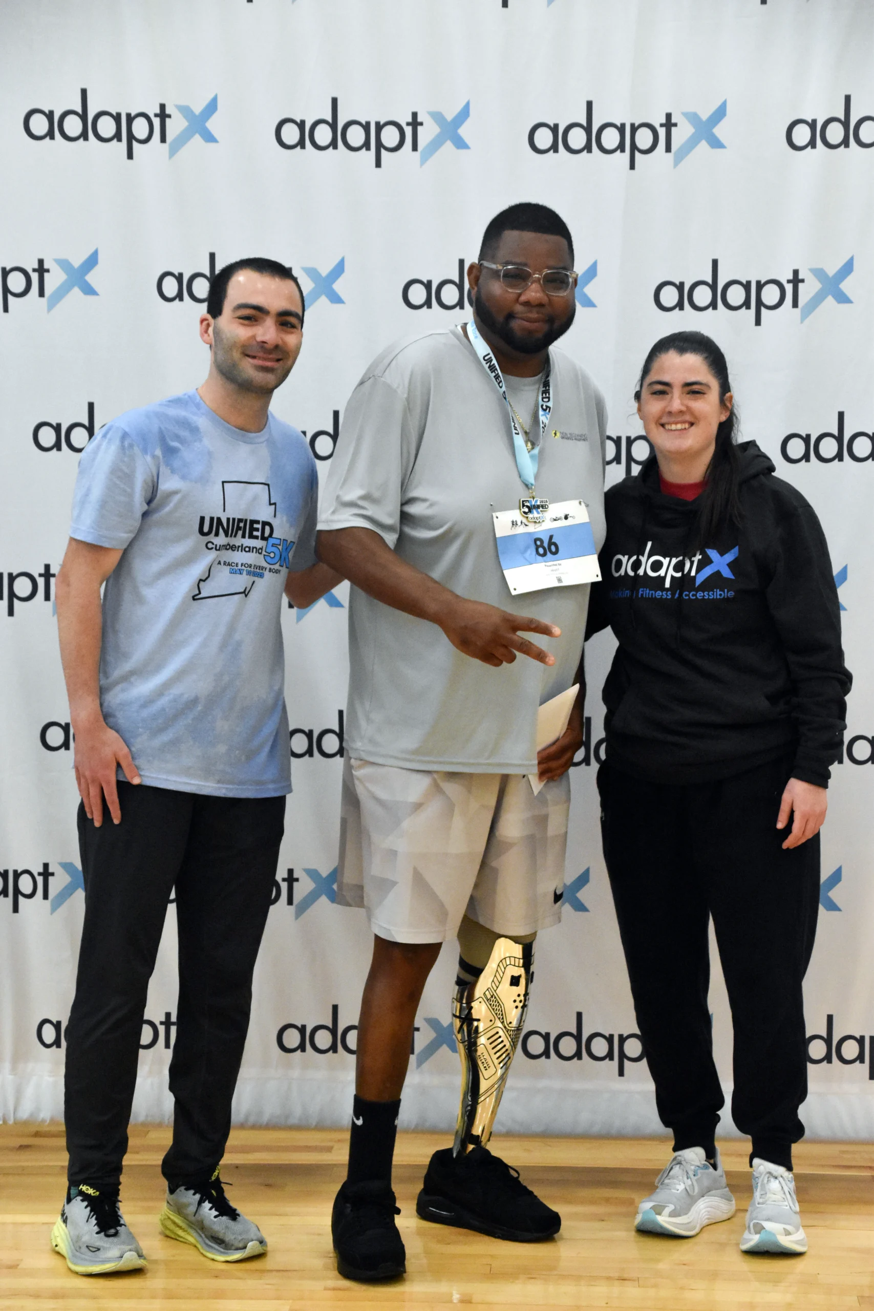 Three people smile for a photo in front of an adaptX banner. The man in the center wears a race medal and bib number 86, and has a prosthetic leg with a gold design.