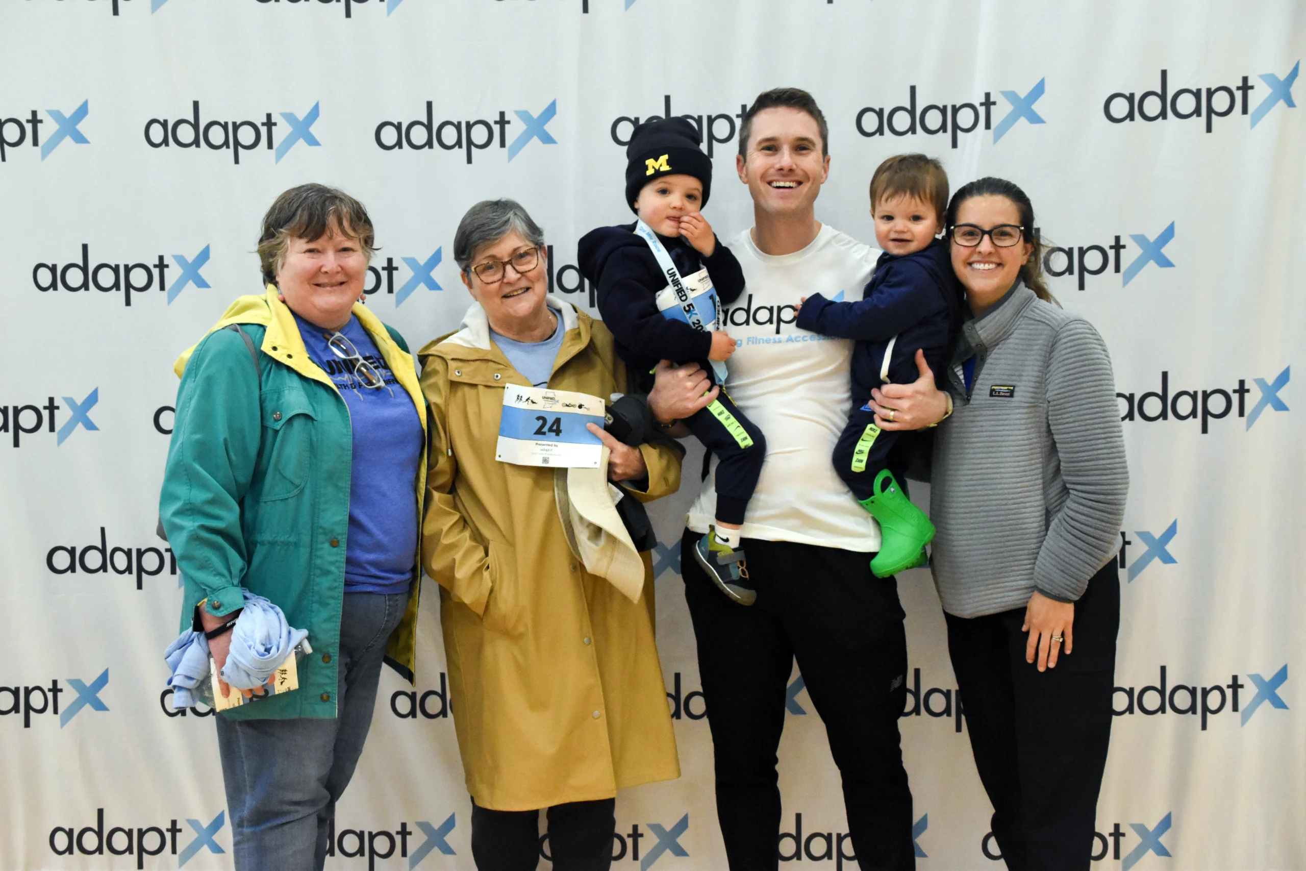 Six people, including a man holding two toddlers, smile together in front of an adaptX step-and-repeat backdrop; one woman wears a race bib and a yellow coat.