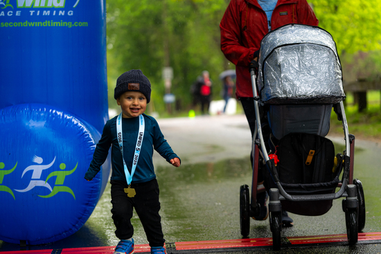 A smiling toddler wearing a medal and beanie walks past the adaptX 5K finish line beside a stroller, with an adult and race arch in the background.