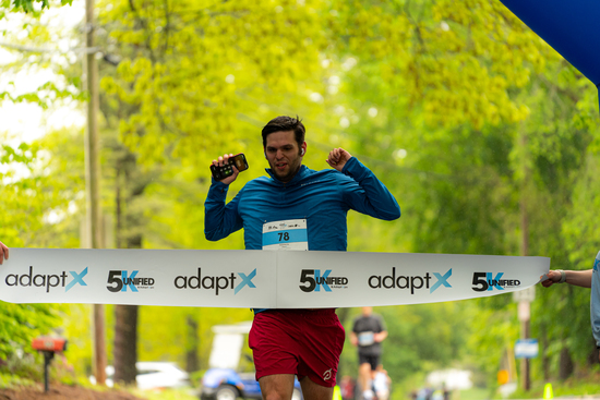 A male runner in a blue shirt and red shorts crosses the adaptX 5K finish line outdoors, raising his arms in celebration while holding a phone.