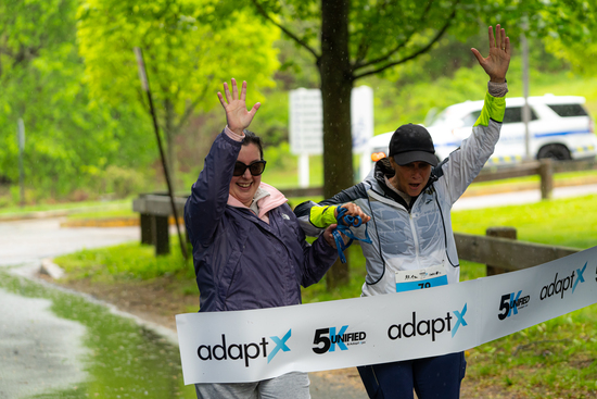 Two women cross the adaptX 5K finish line hand in hand with big smiles and raised arms, celebrating together on a rainy, tree-lined path.