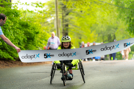 A young athlete in a neon yellow outfit and helmet smiles while crossing the adaptX 5K finish line on a handcycle, surrounded by trees and cheering spectators.