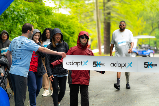 A participant in a red rain jacket and sunglasses crosses the adaptX 5K finish line, surrounded by smiling supporters on a tree-lined road.